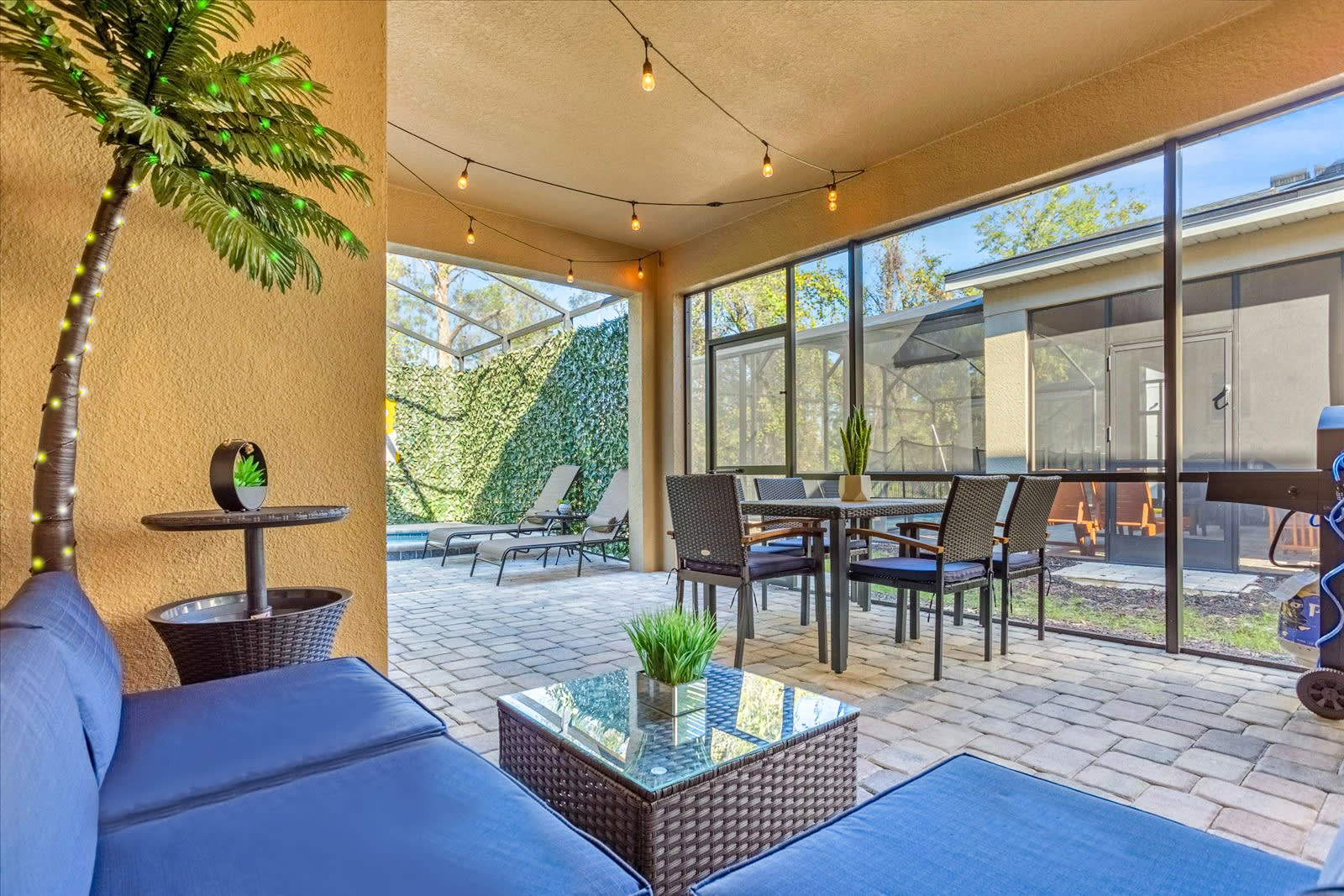 A screened-in patio features a blue sectional sofa, a glass-top coffee table, and a dining set for four, all on light-colored paver flooring. String lights hang overhead, and a decorative palm tree adds a tropical touch, with a pool area visible through the screen.