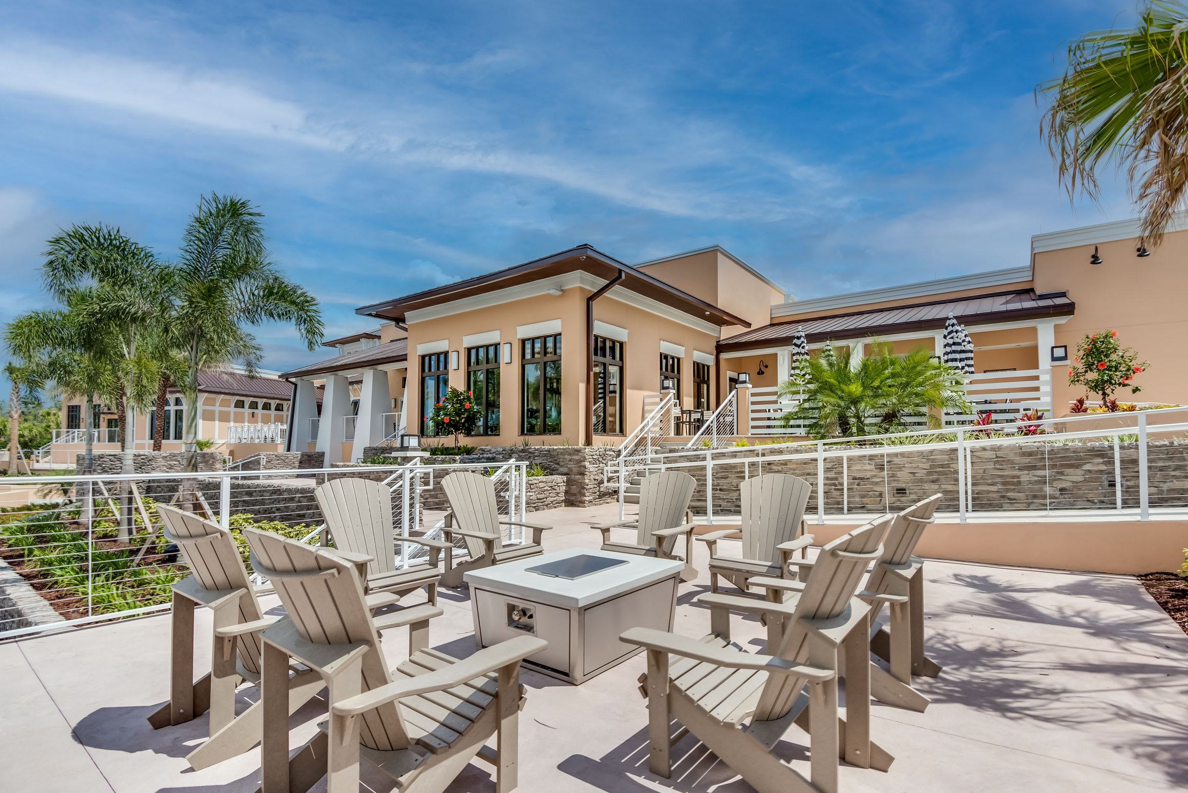 A cozy outdoor fire pit area features a central square fire pit surrounded by several light-colored Adirondack chairs, set on a paved surface with a resort building in the background.