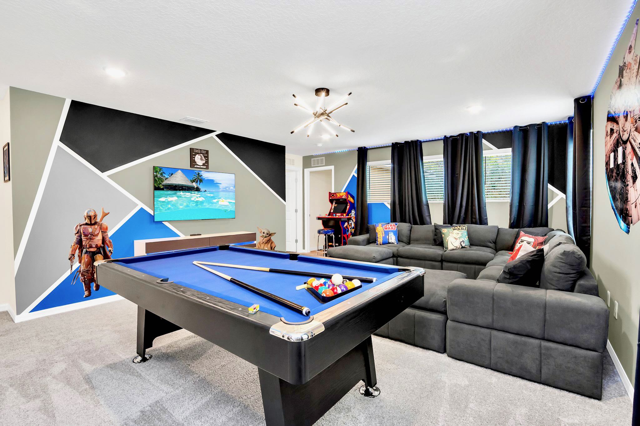 Modern game room with a blue felt pool table, a large gray sectional sofa, a wall-mounted TV, and arcade machines, all set against a geometric black, gray, and blue accent wall.