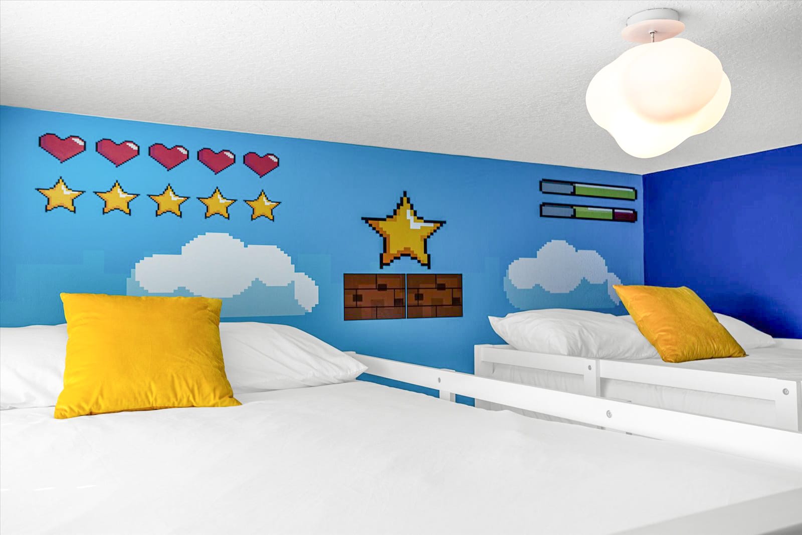 An overhead view of the Super Mario-themed bedroom, showcasing the pixelated wall mural with hearts, stars, and clouds, along with two single beds dressed in white linens and yellow accent pillows. A modern white ceiling light fixture illuminates the room.