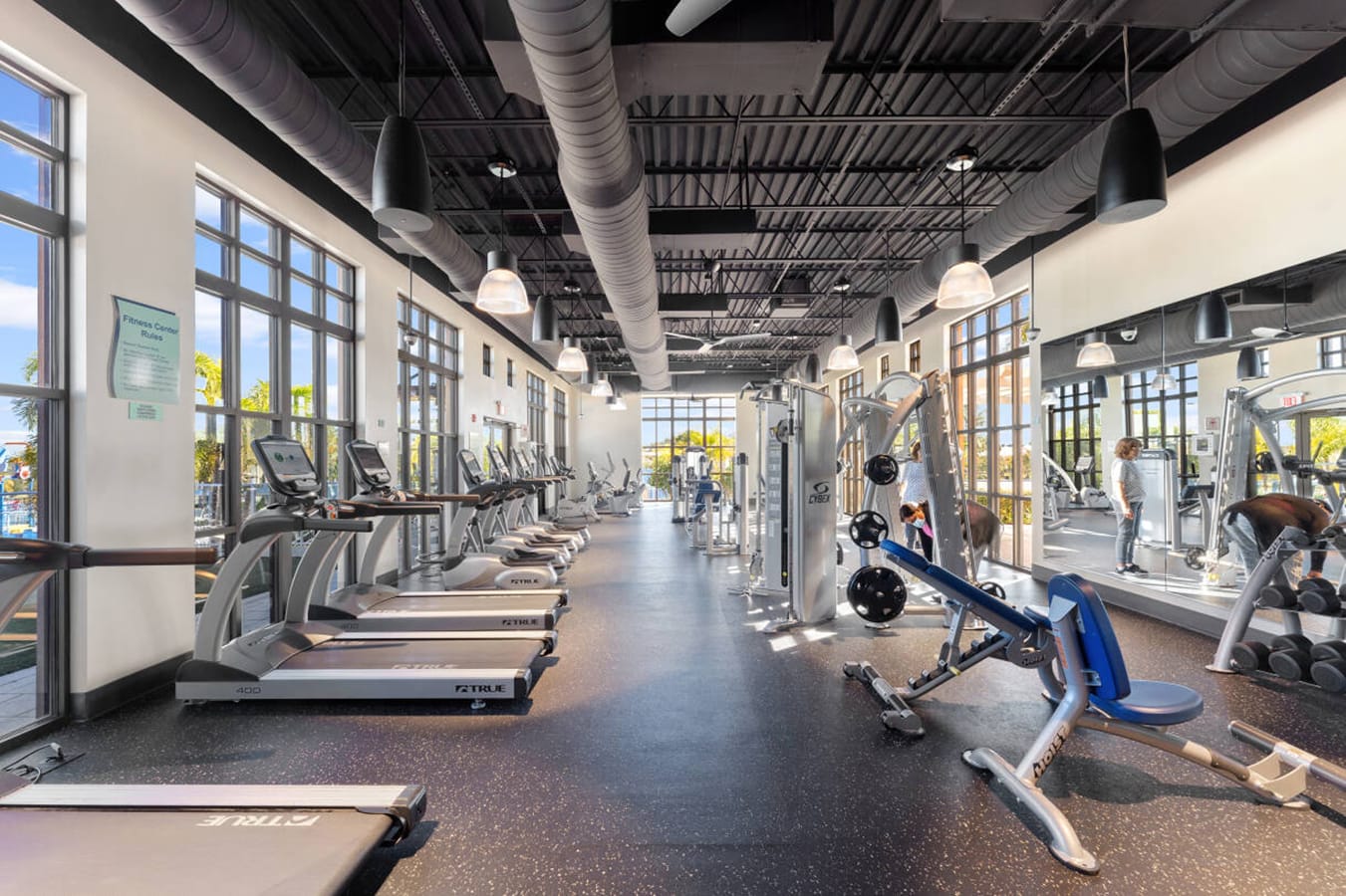 A modern fitness center boasts a wide array of exercise equipment including treadmills, weight machines, and free weights, set on a dark speckled floor under a high exposed ceiling with large windows offering natural light.