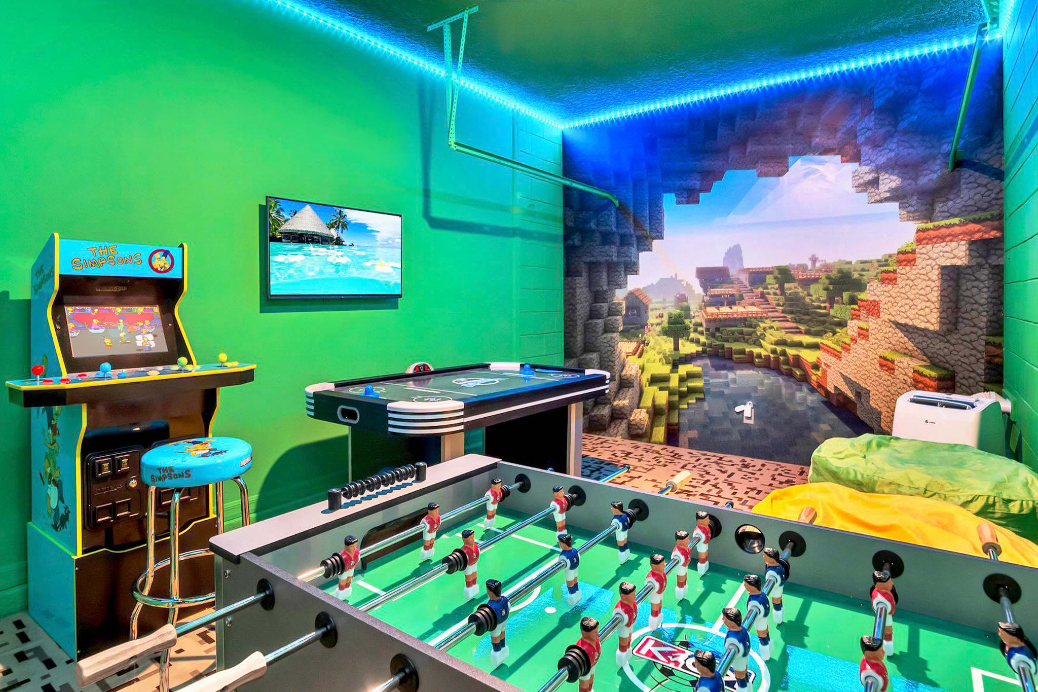 A vibrant game room features bright green walls, blue LED strip lighting, and a large Minecraft-themed mural. It includes a Simpsons arcade machine, a foosball table, and a small pool table, all on a patterned tile floor.