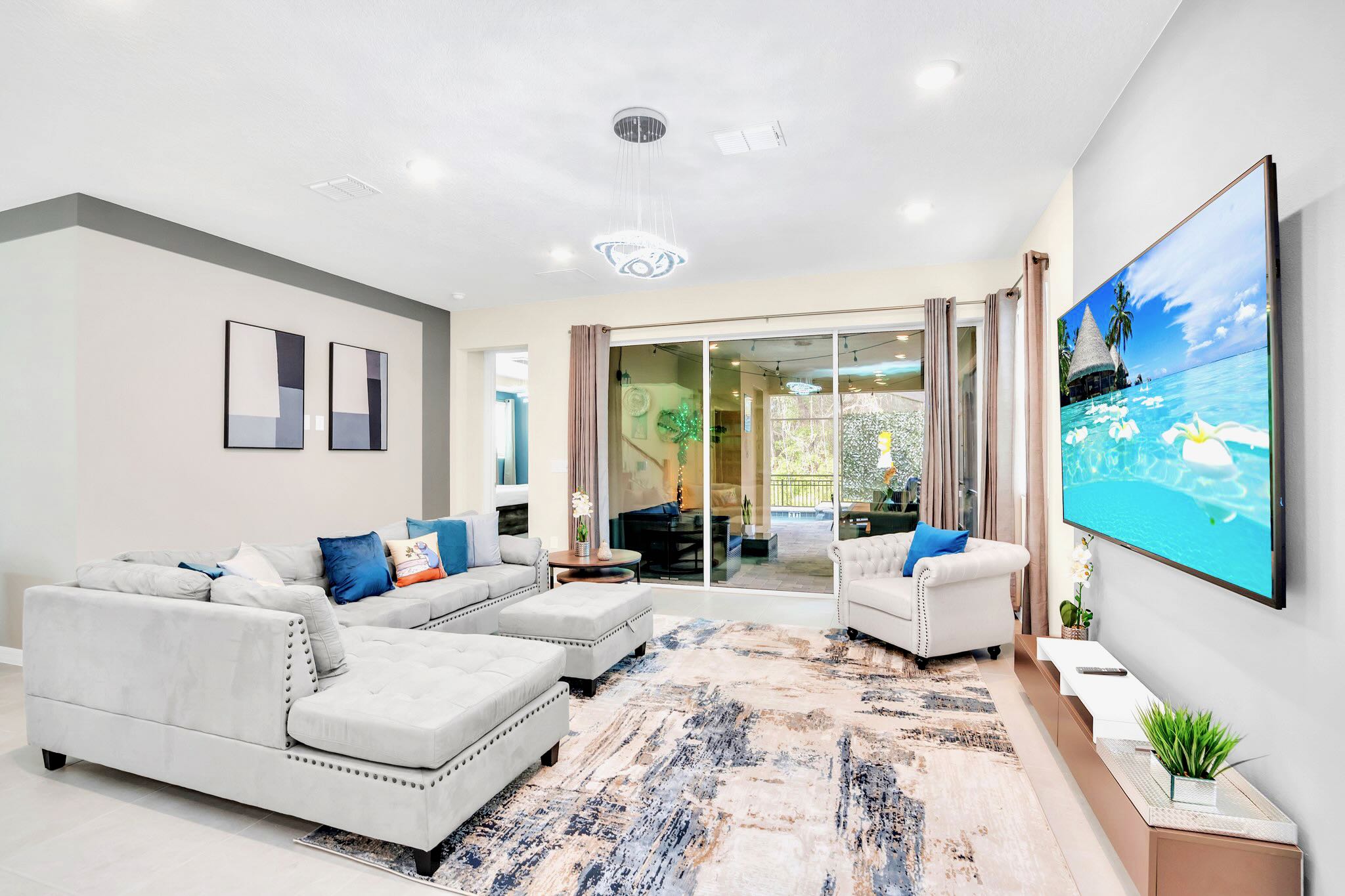 Bright and spacious living room with a light gray sectional sofa, a white armchair, a large wall-mounted TV, and sliding glass doors opening to the screened pool area.
