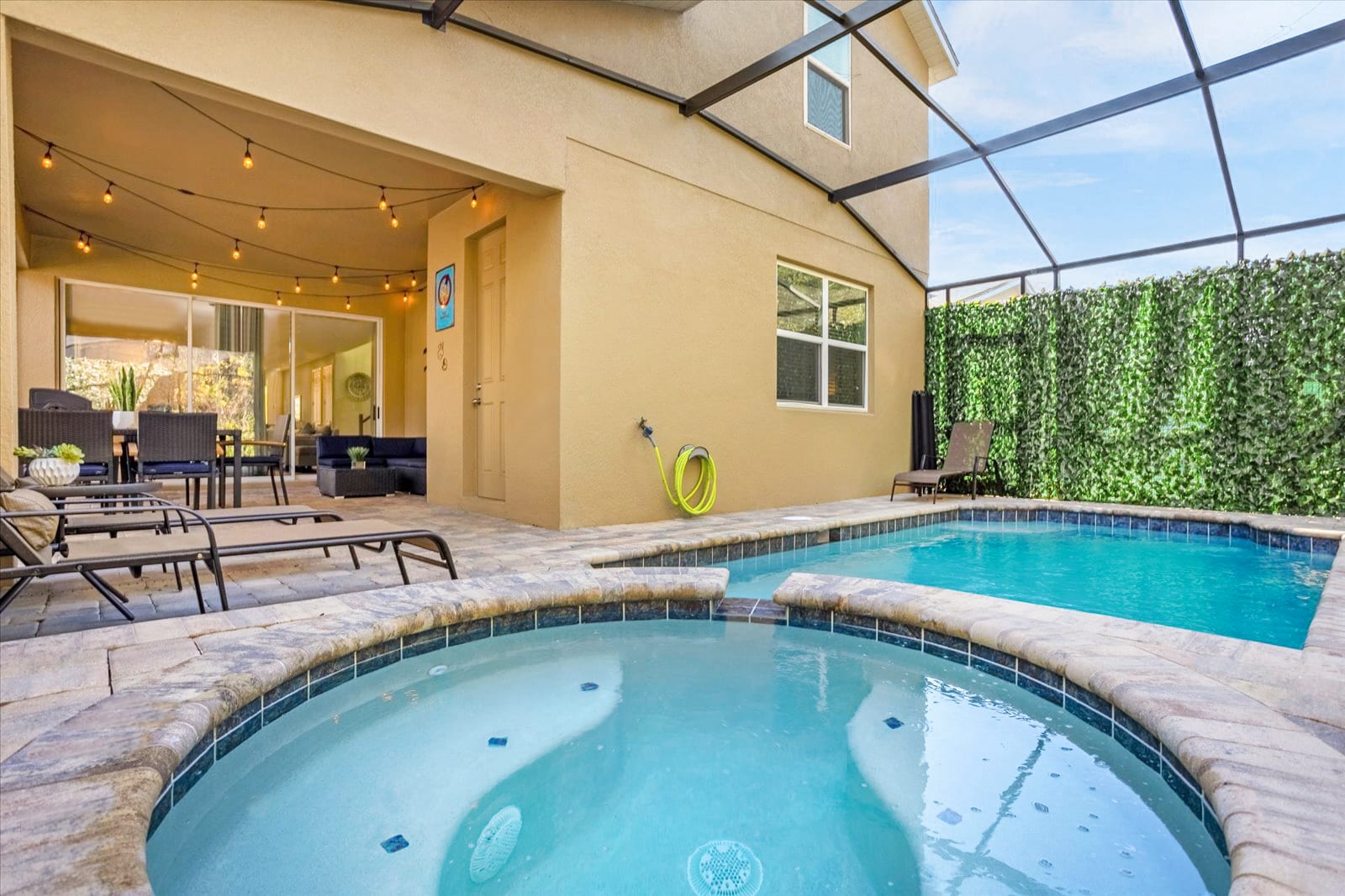 Screened-in outdoor pool and hot tub area with a paved deck, showing the back of the house with a covered patio featuring outdoor seating and string lights.