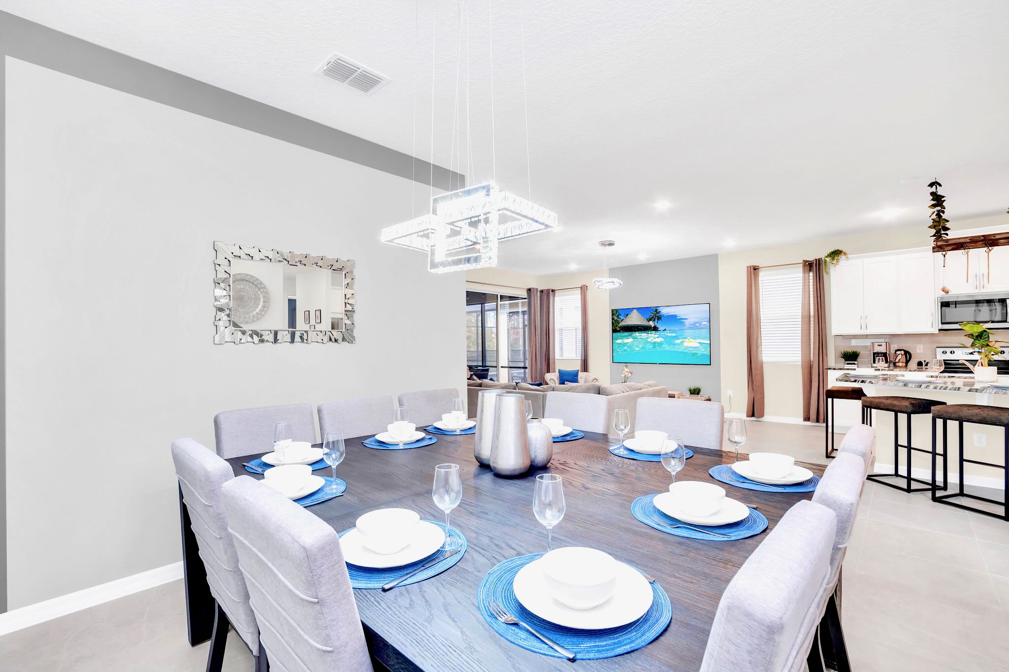 A formal dining area features a long dark wood table set for eight with blue placemats and white dinnerware, illuminated by a modern rectangular chandelier. The space is open to the living room and kitchen, visible in the background.