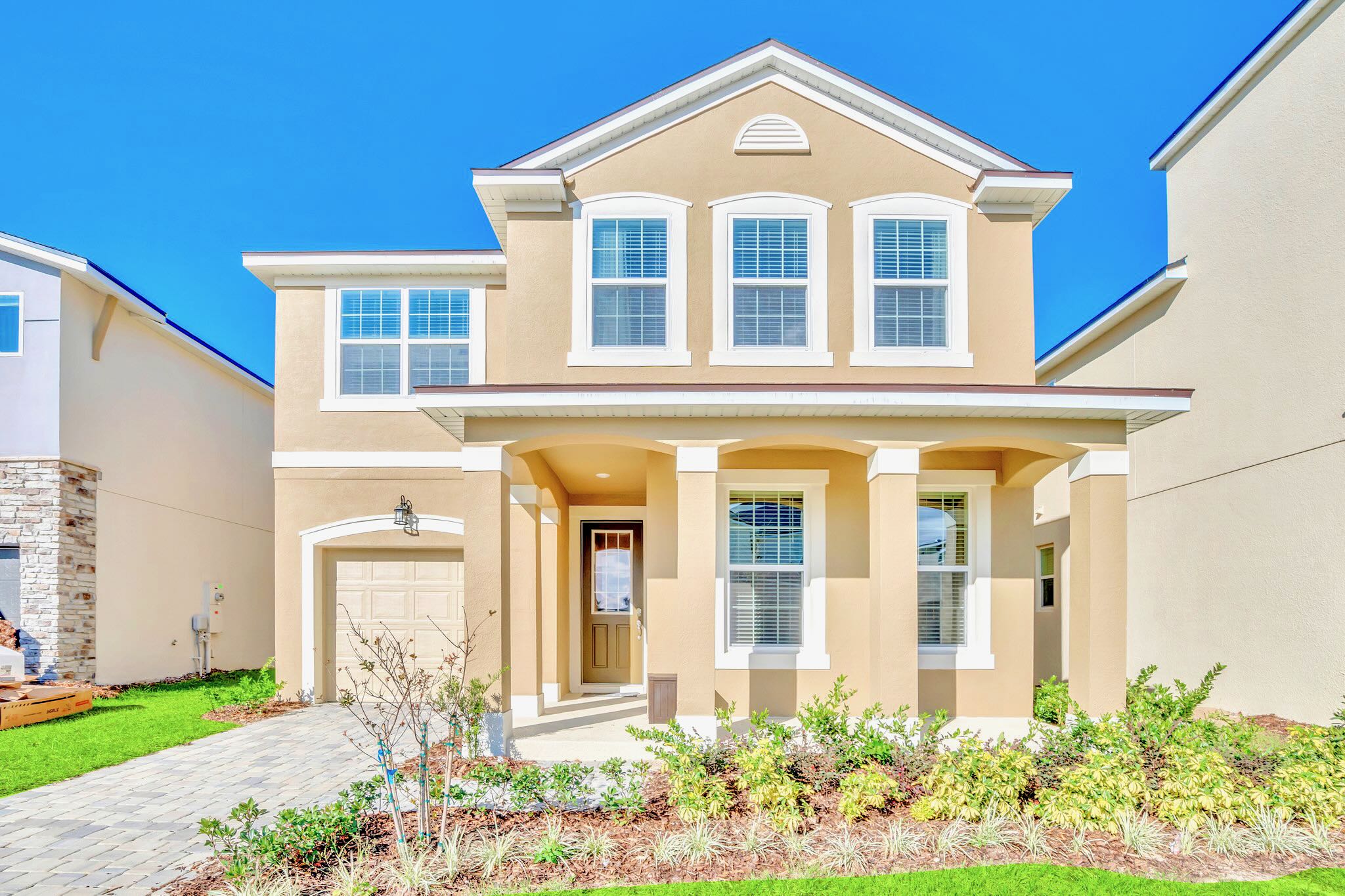 Two-story beige stucco house with white trim, multiple windows, a covered front porch with columns, and a paved driveway leading to a single-car garage.