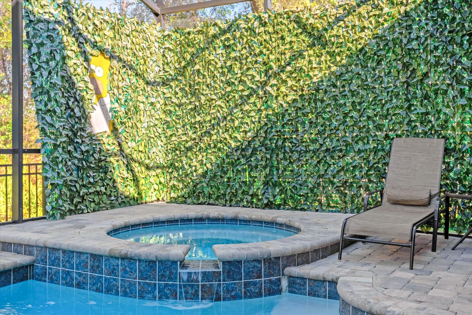 Close-up of the circular hot tub with blue mosaic tiles, surrounded by a stone-edged border and a green faux-foliage wall with a partial view of a yellow character cutout.