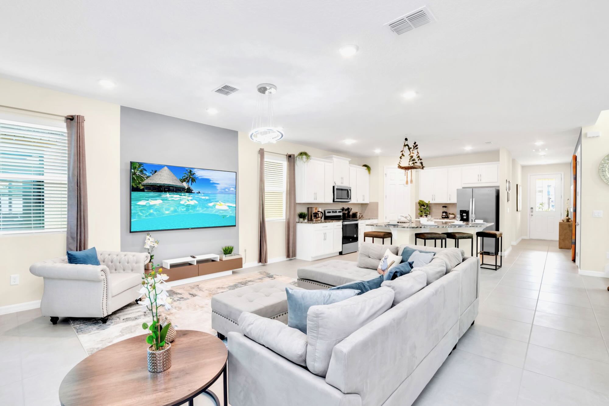 An open-concept living area features a large light gray sectional sofa and a cream armchair, facing a wall-mounted TV displaying a beach scene. The space flows into a modern kitchen with white cabinetry and stainless steel appliances.
