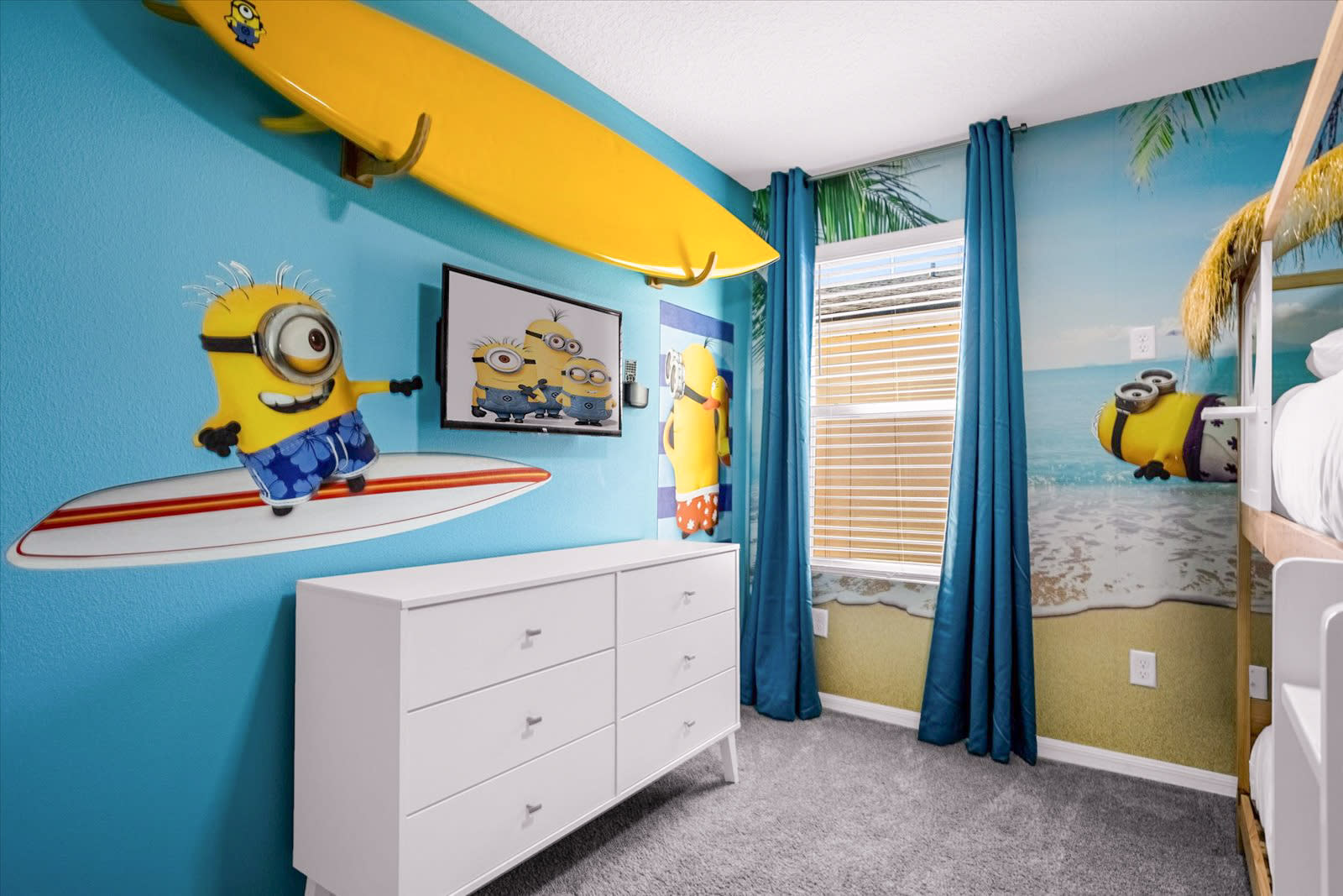 Another view of the Minions-themed bedroom, showcasing a white dresser with six drawers, a wall-mounted TV, and a large yellow surfboard decoratively mounted above the TV. Minion characters are prominently featured on the bright blue and beach-themed wall murals.