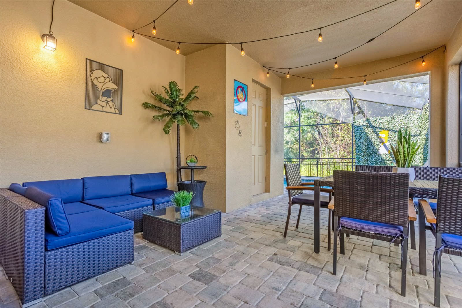 Covered outdoor patio with a blue wicker sectional sofa, a dining table with chairs, and string lights, overlooking the screened pool area.