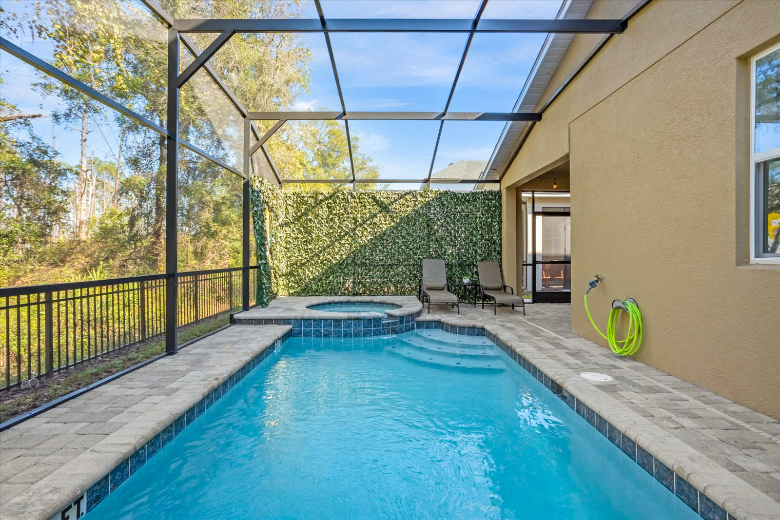 Long, rectangular screened-in private pool with an integrated hot tub, surrounded by a paved deck and a green faux-foliage wall, with lounge chairs.
