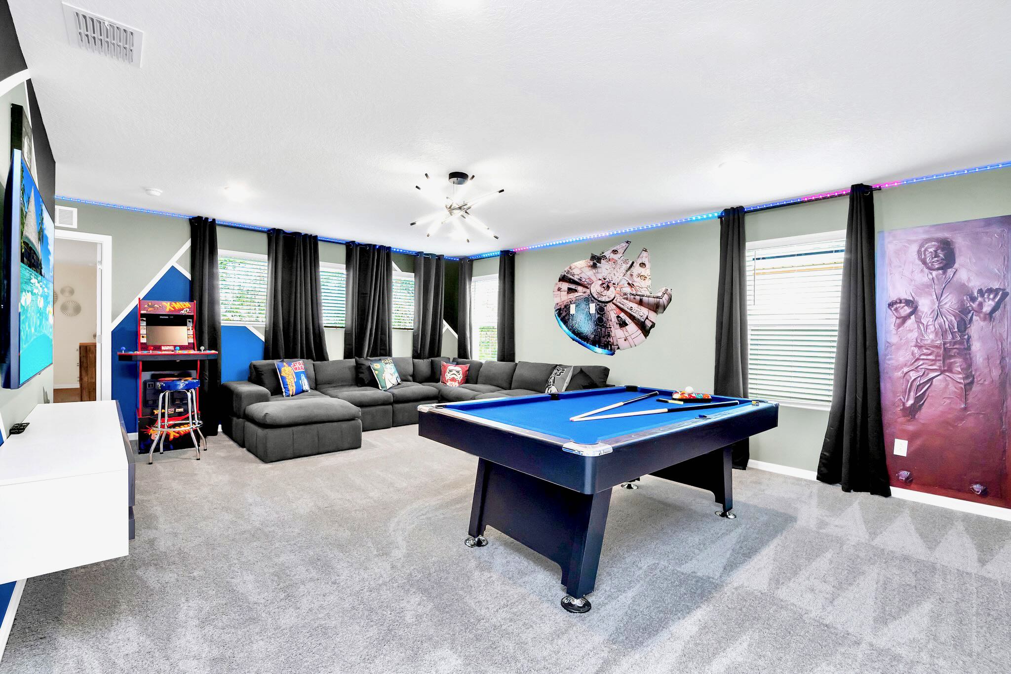 A spacious game room features a large dark gray sectional sofa, a blue felt pool table, and Star Wars-themed decor including a Millennium Falcon wall art and a Han Solo in carbonite painting. The room has light gray walls, dark curtains, and colorful LED strip lighting.