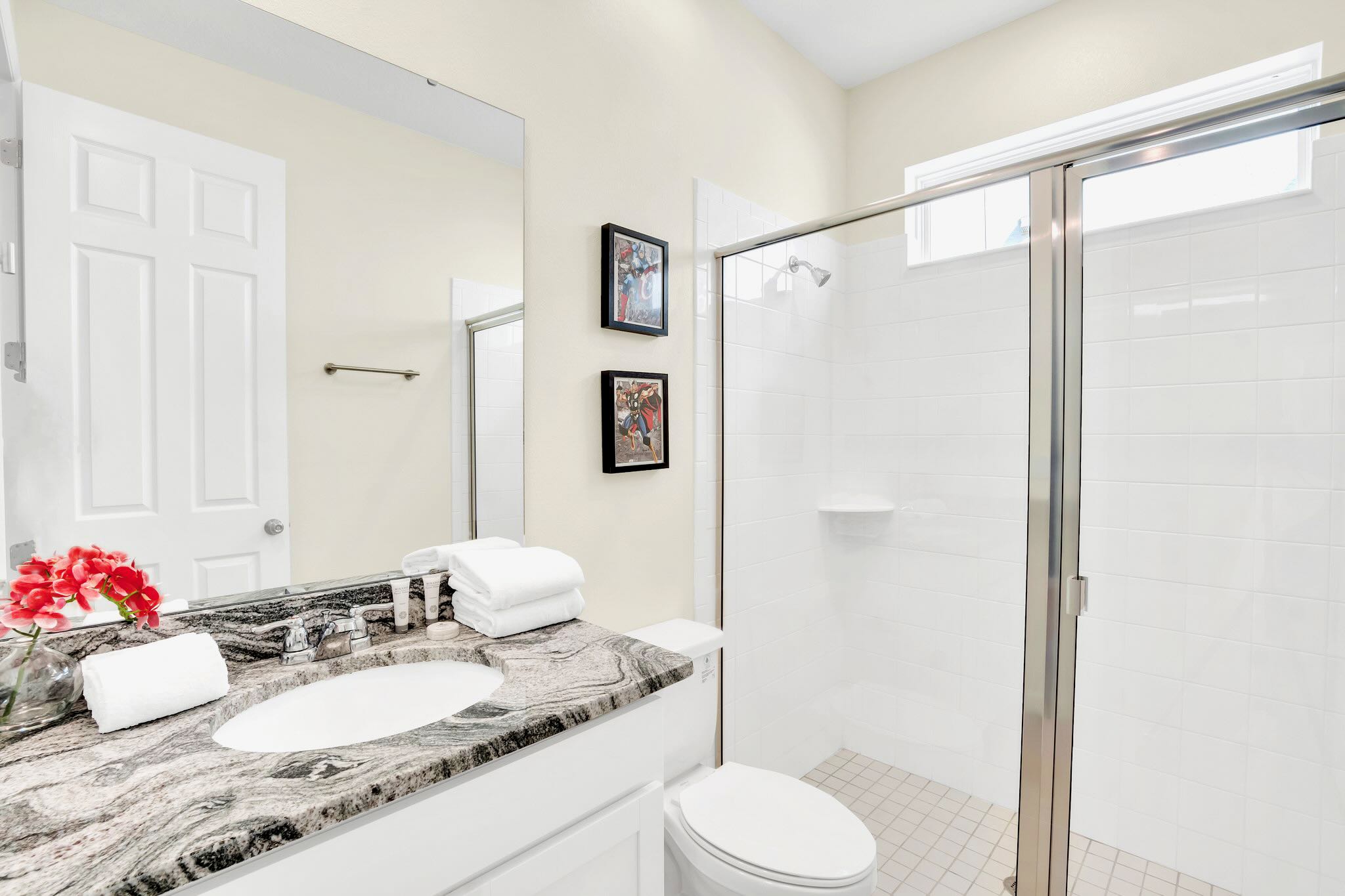A clean bathroom features a single granite vanity with a white sink, a toilet, and a glass-enclosed shower with white tiled walls. Red floral decor adds a pop of color.