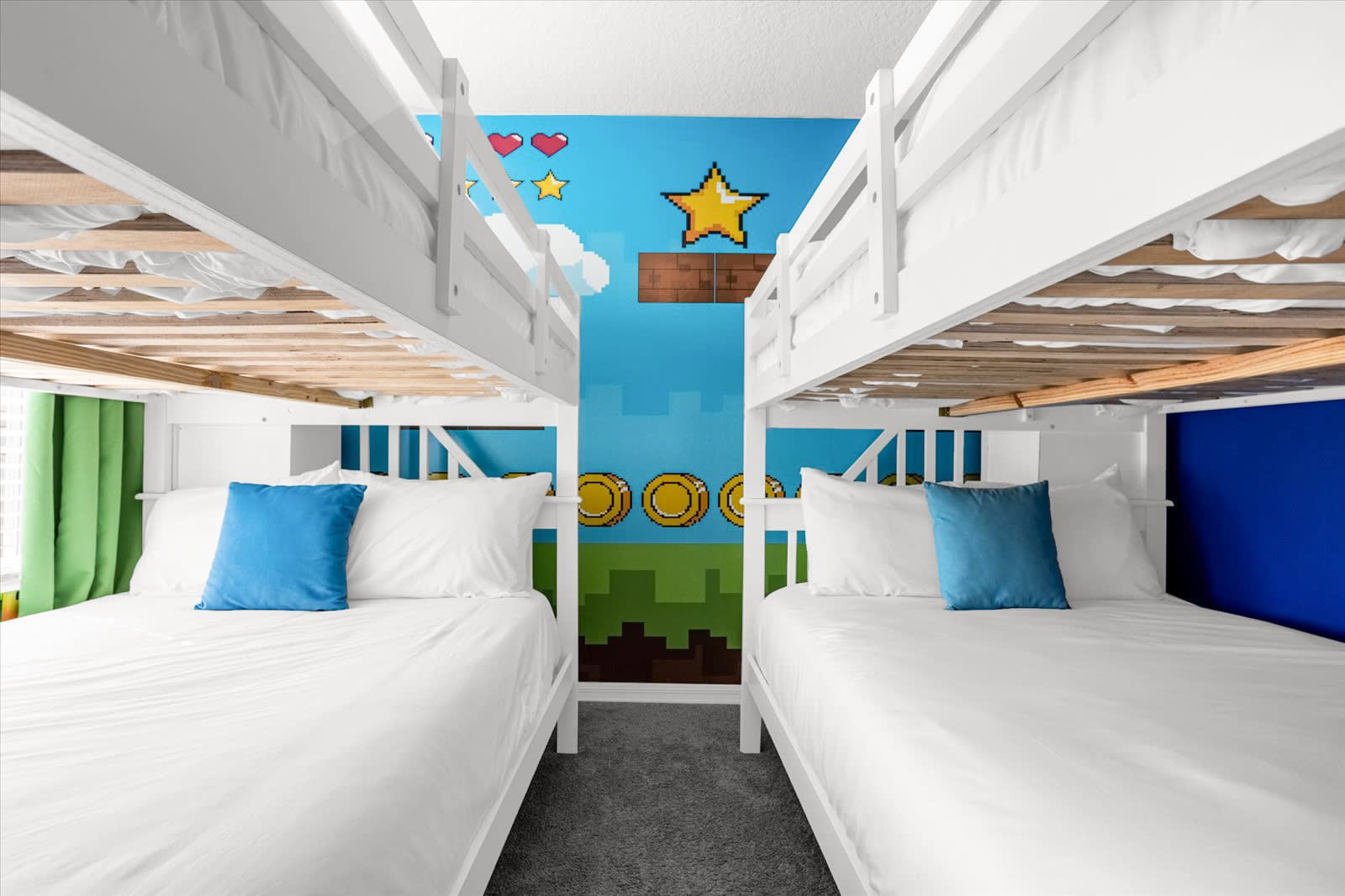 A close-up view between two white wooden bunk beds in the Super Mario-themed bedroom, revealing a pixelated wall mural with classic game elements like coins and green landscape. Each bed is made with white linens and a blue accent pillow.