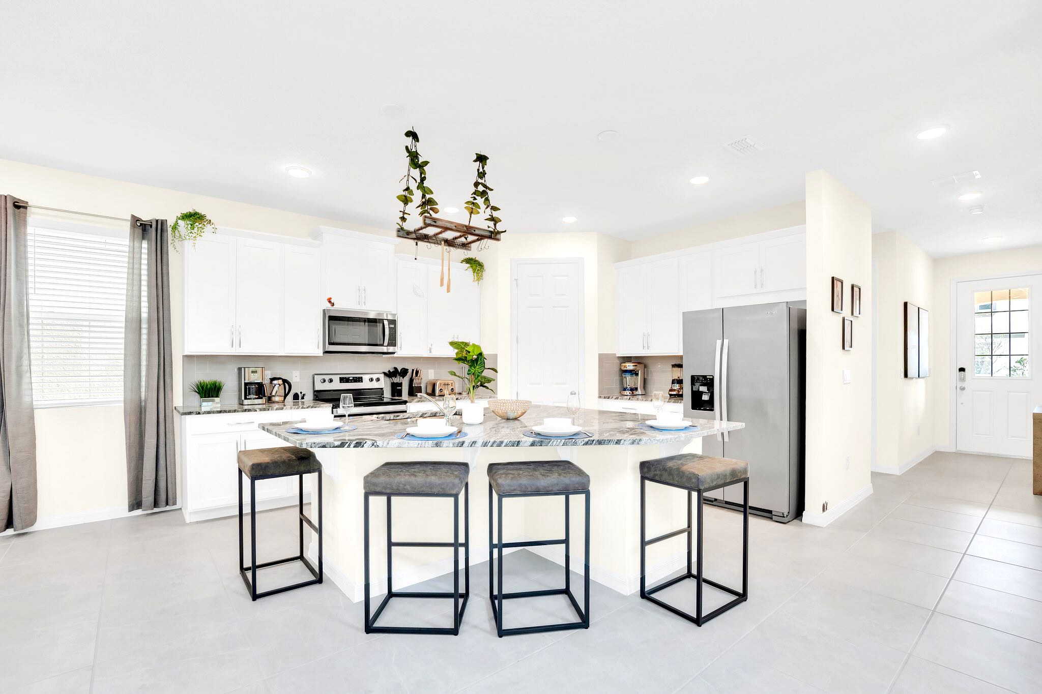 A bright kitchen showcases white cabinetry, granite countertops, and a large island with four dark gray bar stools, serving as a casual dining area. Stainless steel appliances and light tile flooring contribute to the modern aesthetic.