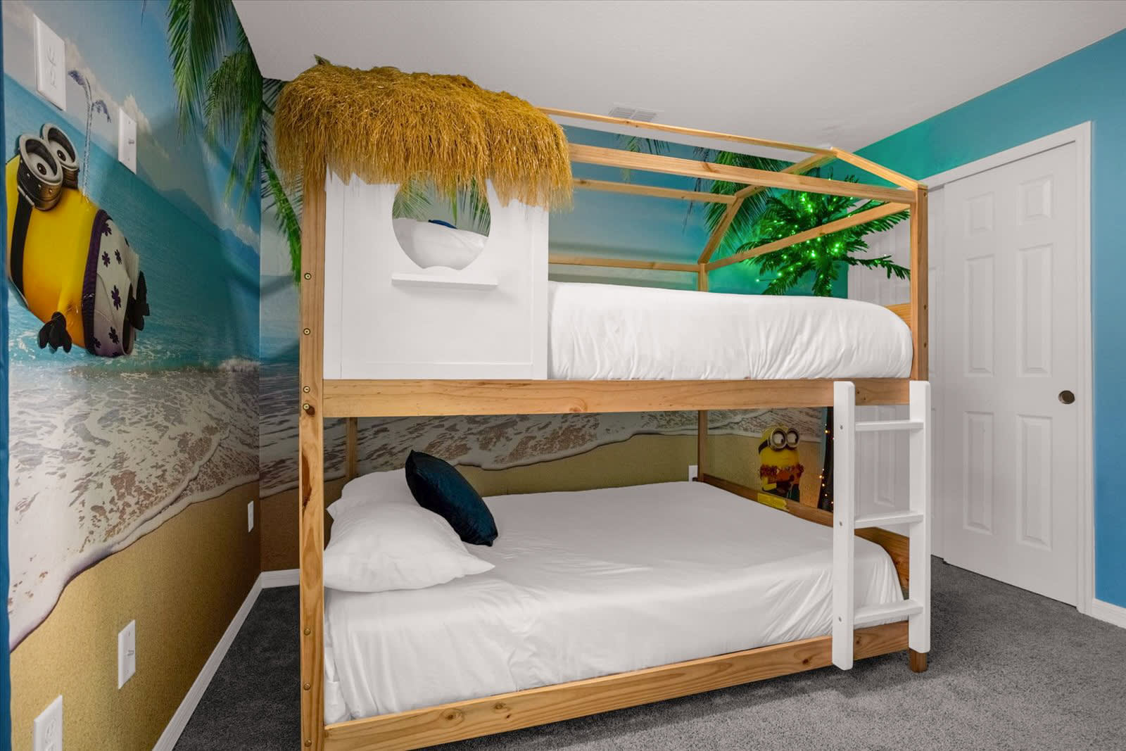 A vibrant Minions-themed bedroom featuring a custom wooden bunk bed designed like a beach hut with a thatched roof, set against a wall mural depicting a tropical beach scene with Minions. The beds are dressed in crisp white linens, and a large Minion character is affixed to the wall.