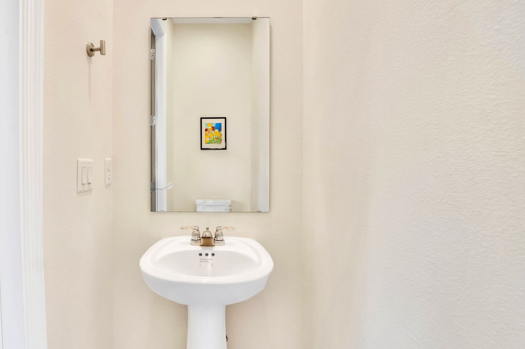 A simple and clean half-bathroom featuring a white pedestal sink with a chrome faucet and a rectangular mirror above it. The walls are painted a light cream color, and a small framed picture adds a subtle decorative touch.