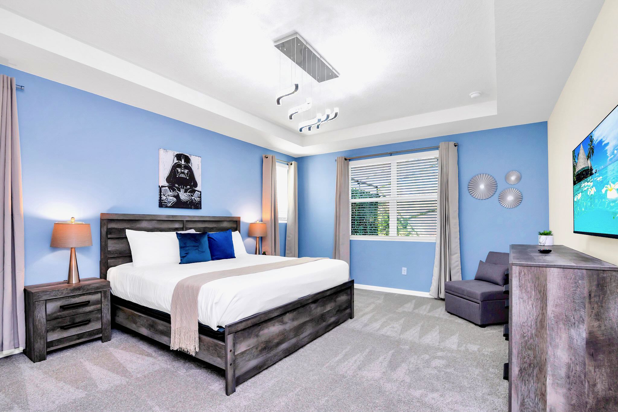 A spacious bedroom with light blue walls features a dark wood king-sized bed with blue accents, two matching nightstands, and a wall-mounted TV. A modern chandelier hangs from the tray ceiling.