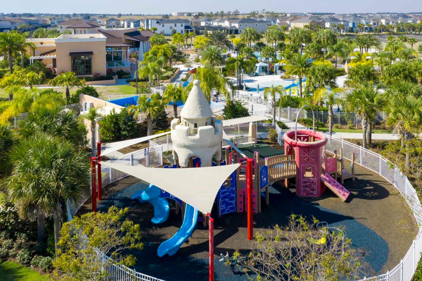 An aerial shot reveals a vibrant children's playground featuring a unique sandcastle-themed play structure, multiple slides, and shaded areas, all enclosed by a white fence within a lush green landscape.