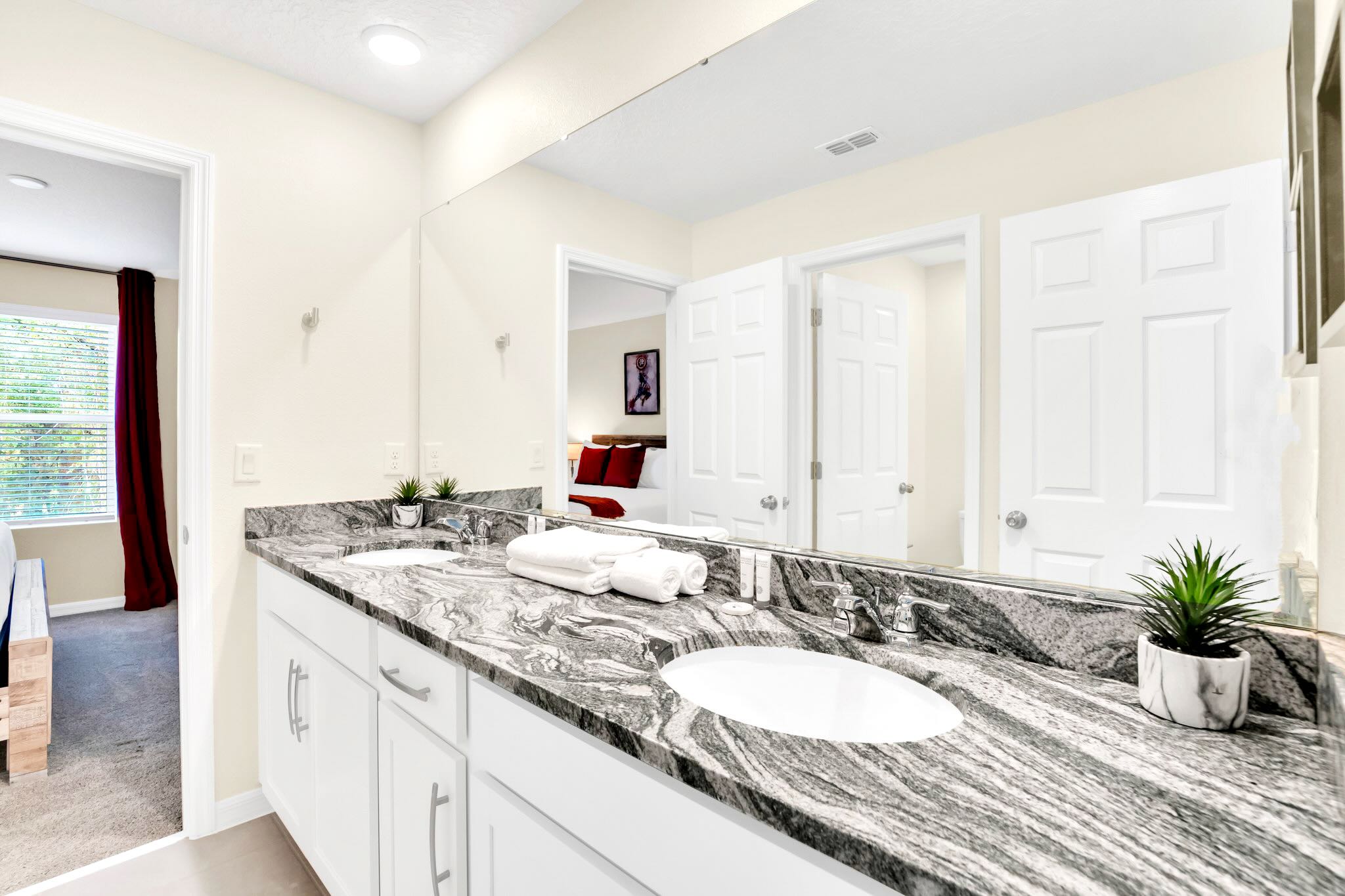 A spacious bathroom features a double vanity with a dark granite countertop and two white sinks, a large mirror, and white cabinets. The reflection shows a bedroom with a wooden bed and red accents.