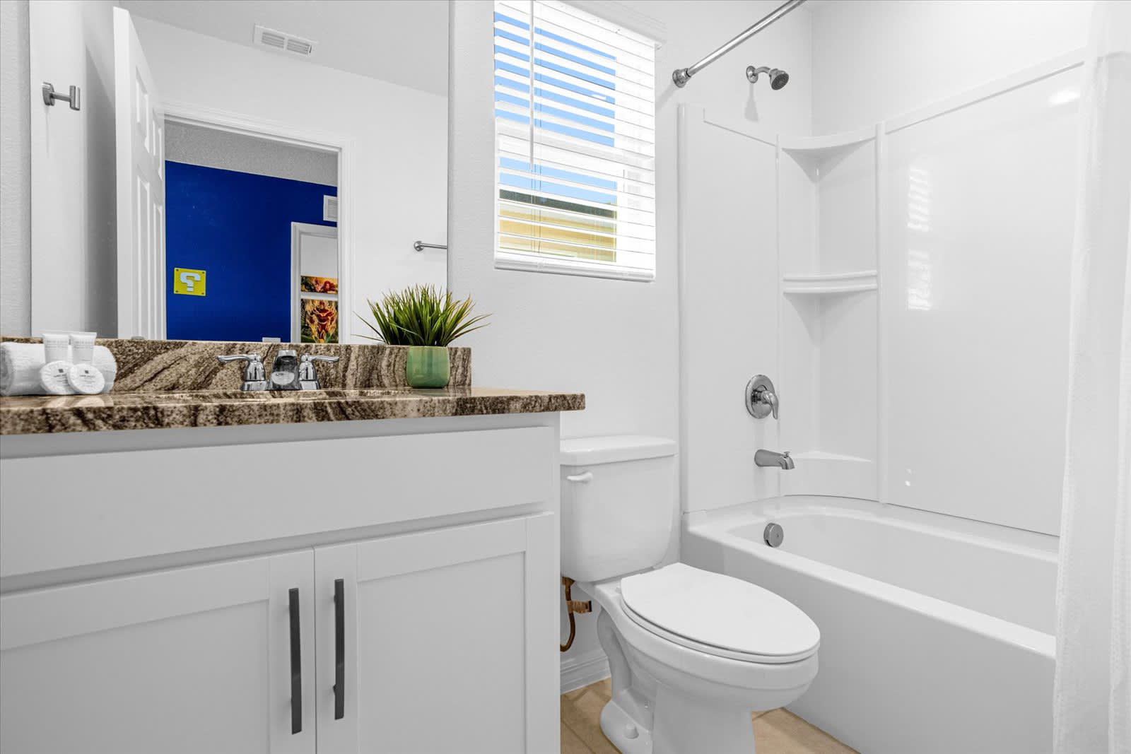 A bright bathroom featuring a white vanity with a granite countertop and a single sink, a toilet, and a white shower/bathtub combo with a chrome fixture. A window with white blinds provides natural light, and a small green plant adds a touch of decor.