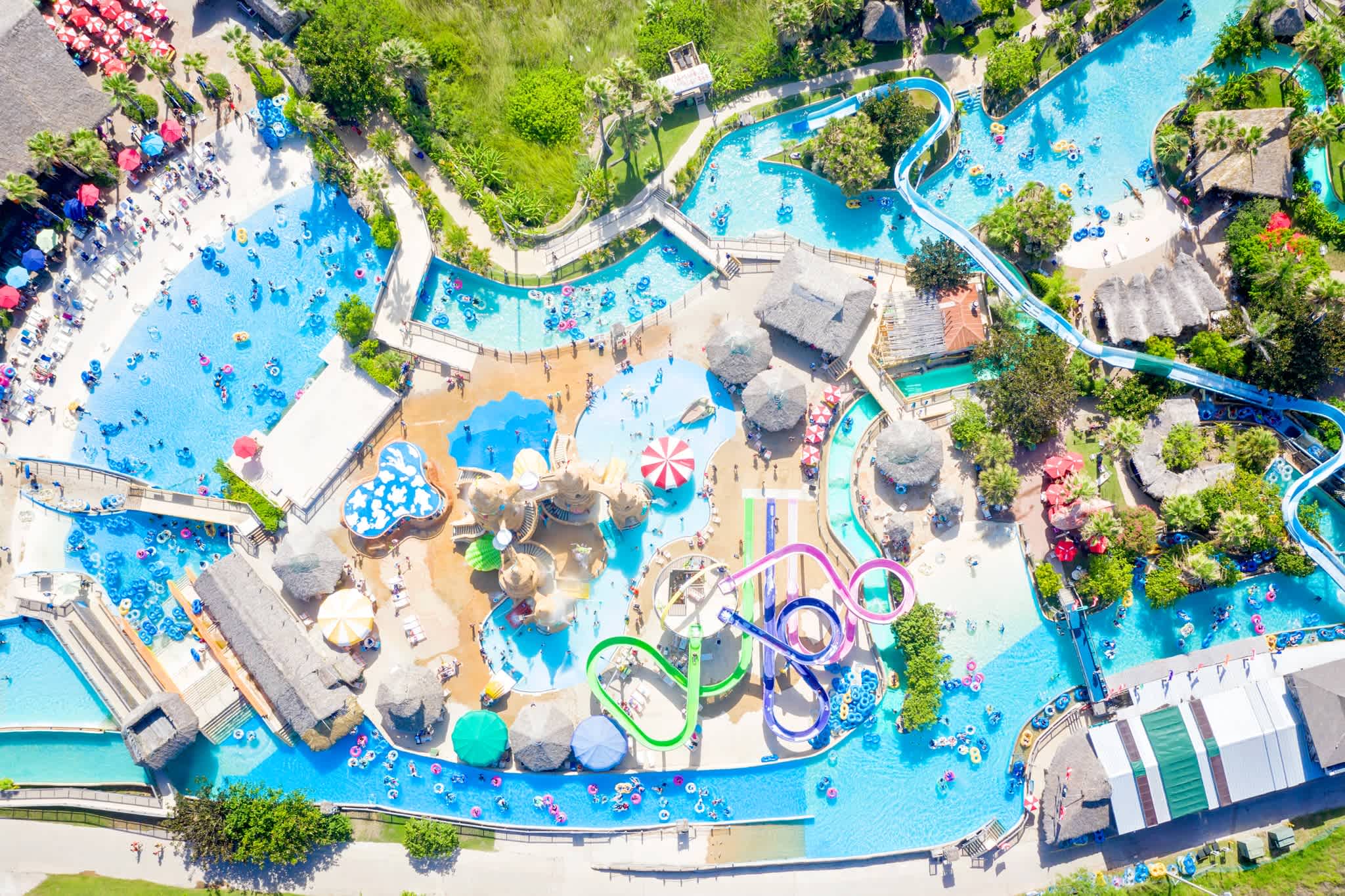Aerial view of a vibrant water park featuring multiple interconnected pools, winding lazy rivers, colorful water slides, and numerous guests enjoying the various attractions under a bright sky.