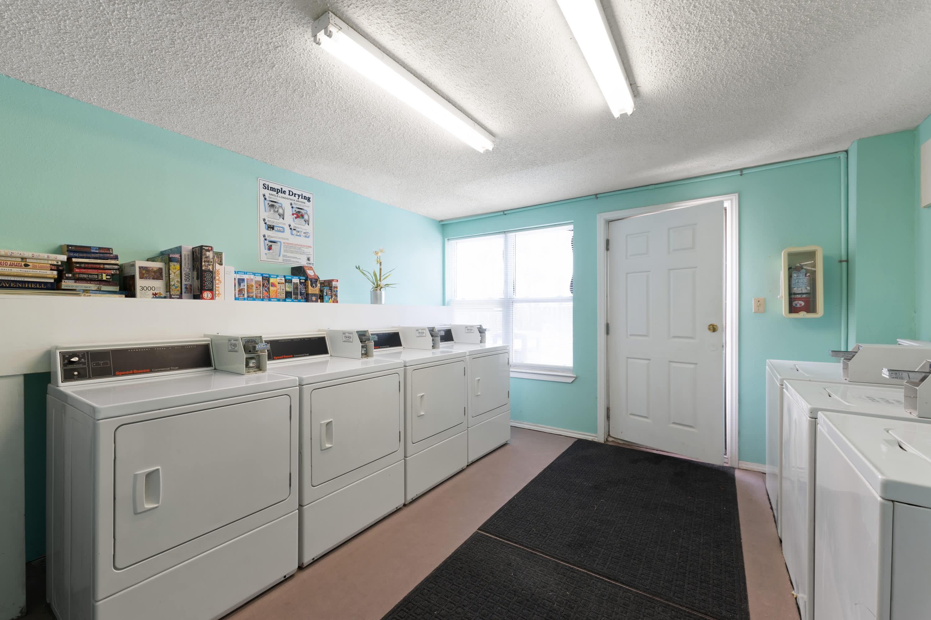 A communal laundry room with several white top-loading washing machines and front-loading dryers. The walls are painted a bright teal, and a window provides natural light.