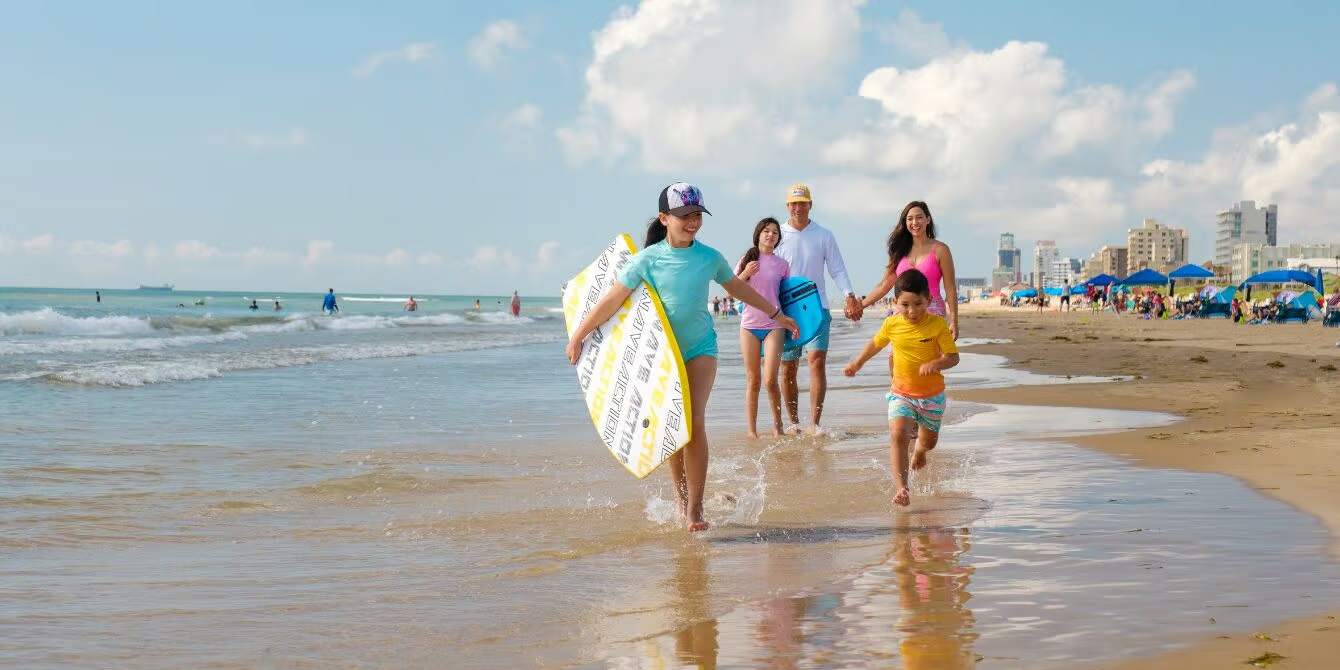 A joyful family of four, including two adults and two children, runs out of the clear ocean water onto a wide sandy beach, with one child carrying a boogie board and resort buildings visible in the distance.