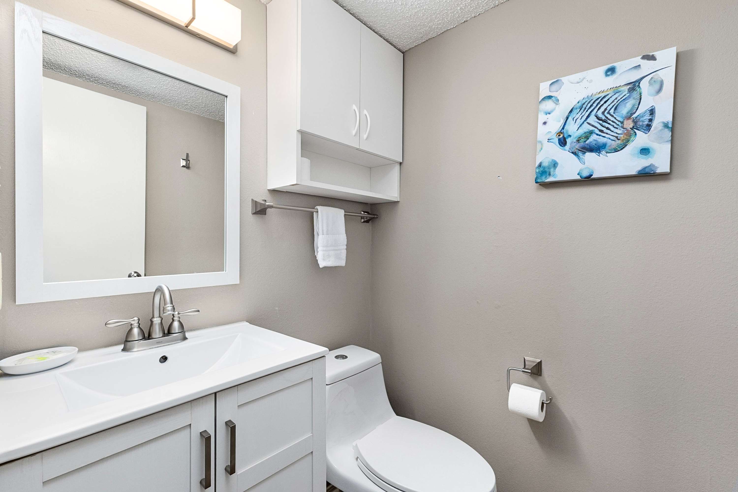 A compact bathroom with a white vanity and sink, a toilet, and a mirror. White wall cabinets provide storage, and a blue fish artwork adds a coastal touch.