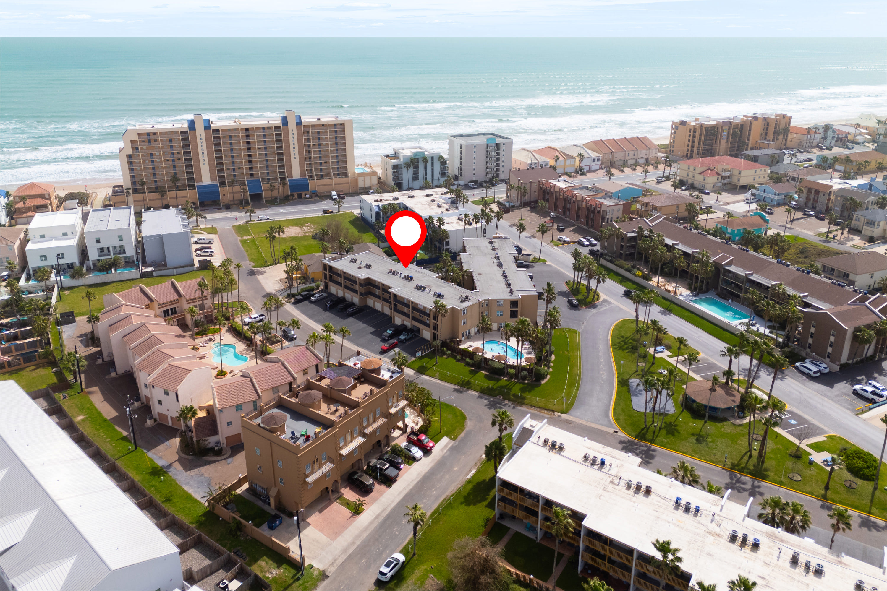 An aerial view of the condominium complex, showing multiple buildings, parking lots, and proximity to the beach and ocean. A red map pin highlights the property location.