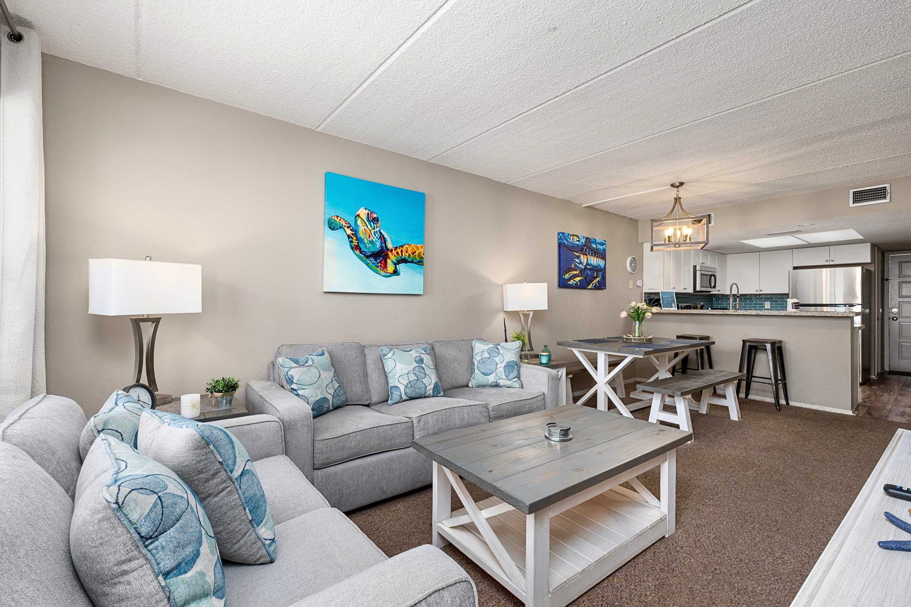 An open-concept living and dining area features a gray sofa with blue patterned pillows, a rustic coffee table, and a dining table with benches. Coastal artwork adorns the walls, and the space flows into the kitchen area visible in the background.