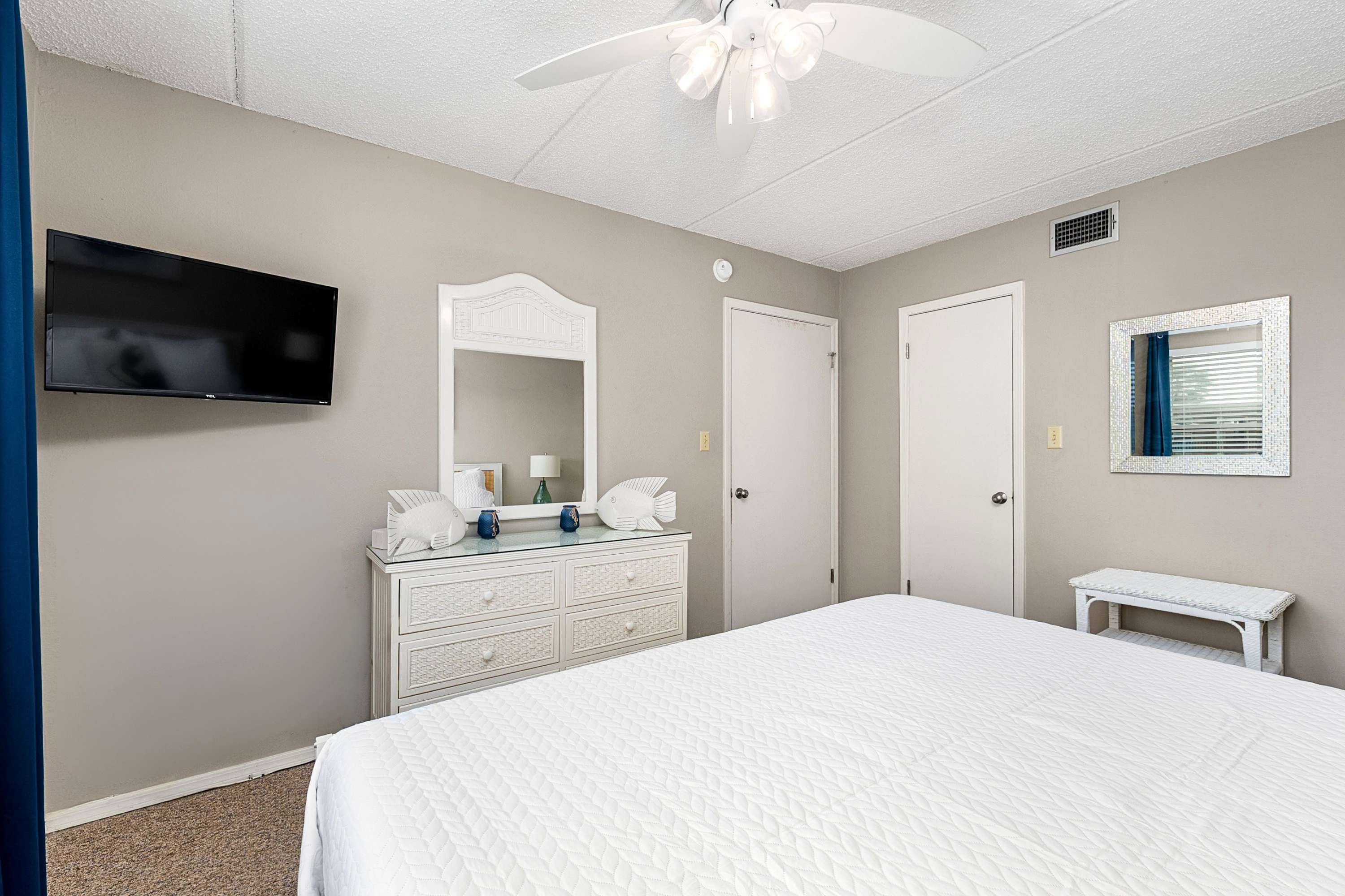 Another view of the bedroom, featuring a white wicker dresser with a mirror, a wall-mounted TV, and two white doors. Decorative white fish sculptures adorn the dresser.