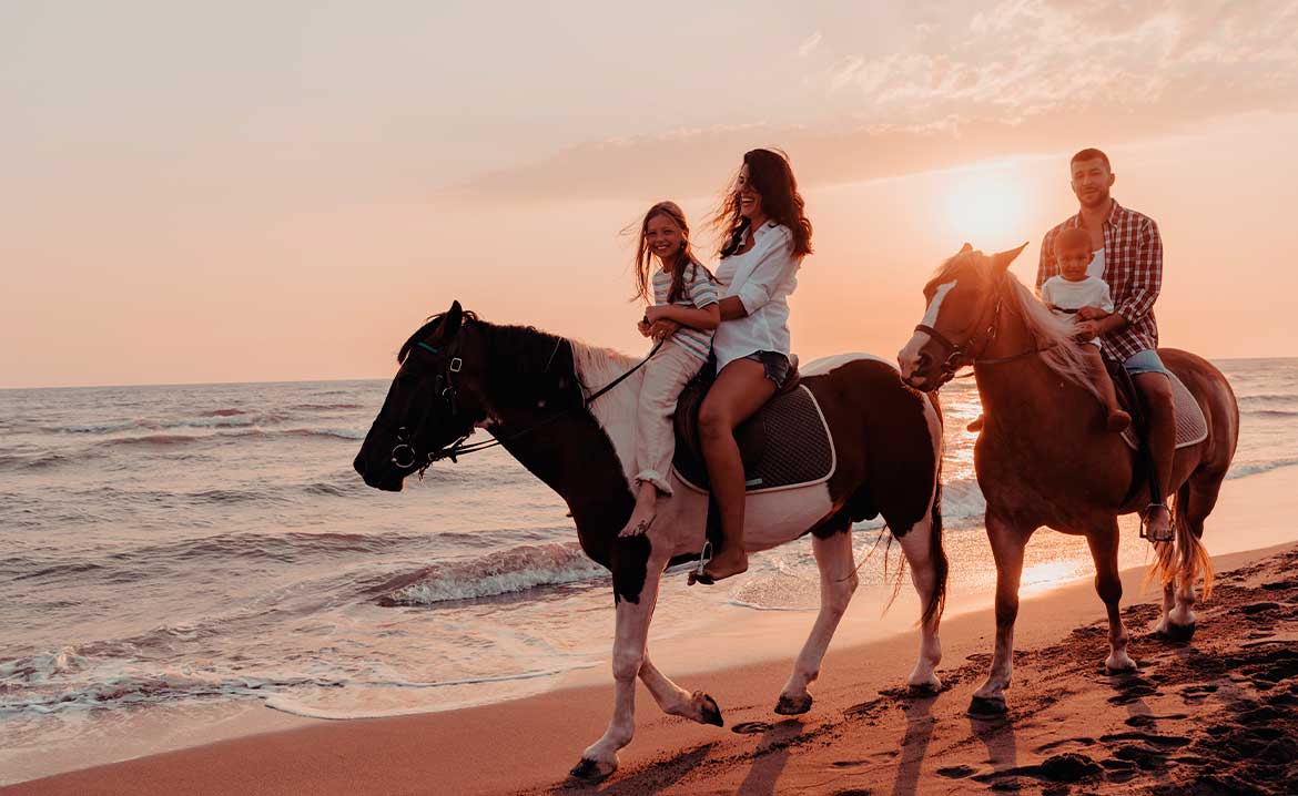 A family of four, including two adults and two children, enjoys a scenic horseback ride along a sandy beach at sunset, with the ocean waves gently lapping the shore in the background.