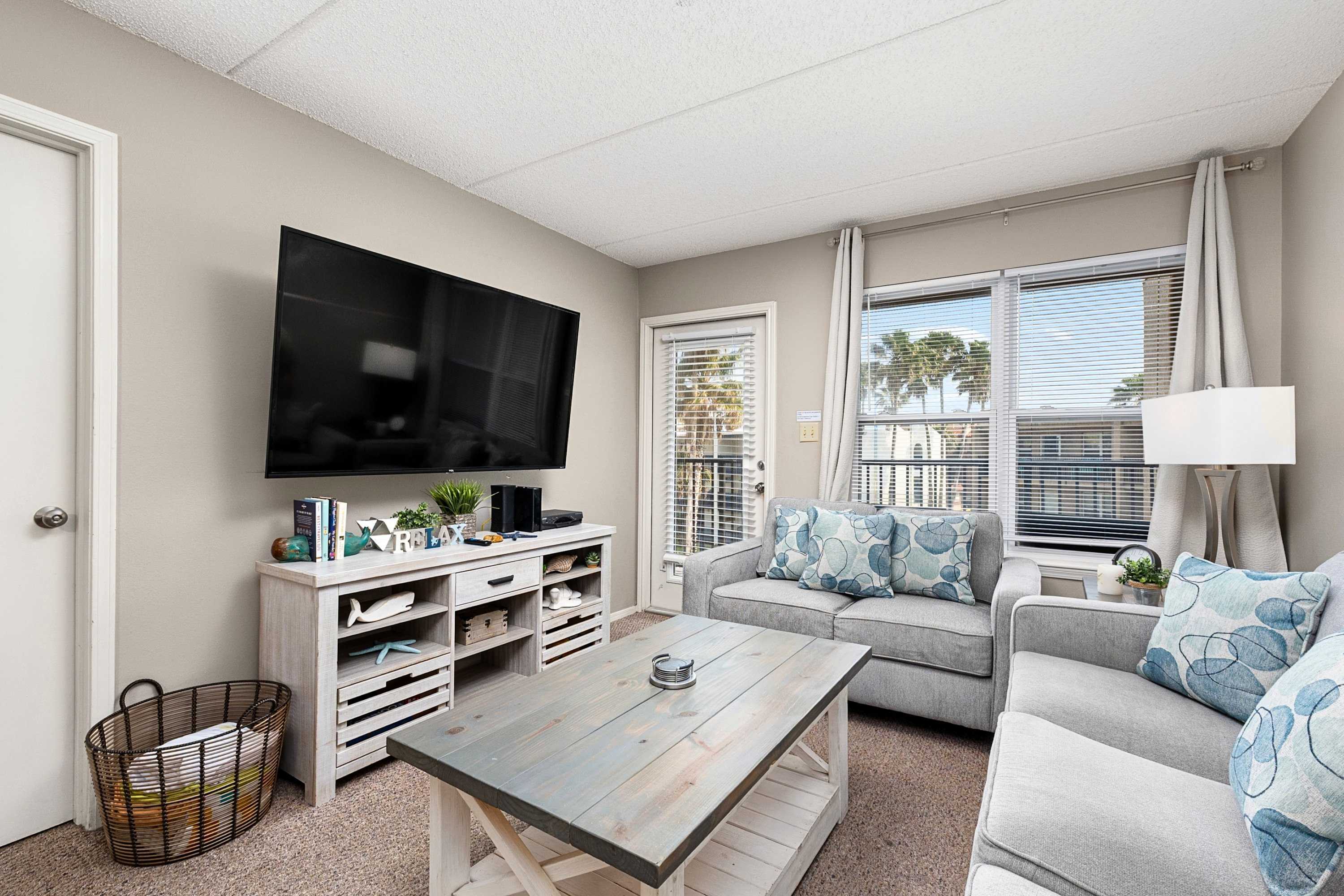 A bright living room features a wall-mounted flat-screen TV above a distressed white media console, a gray armchair, and a rustic white and gray coffee table. A glass door and window offer views outside, and a woven basket sits on the carpeted floor.