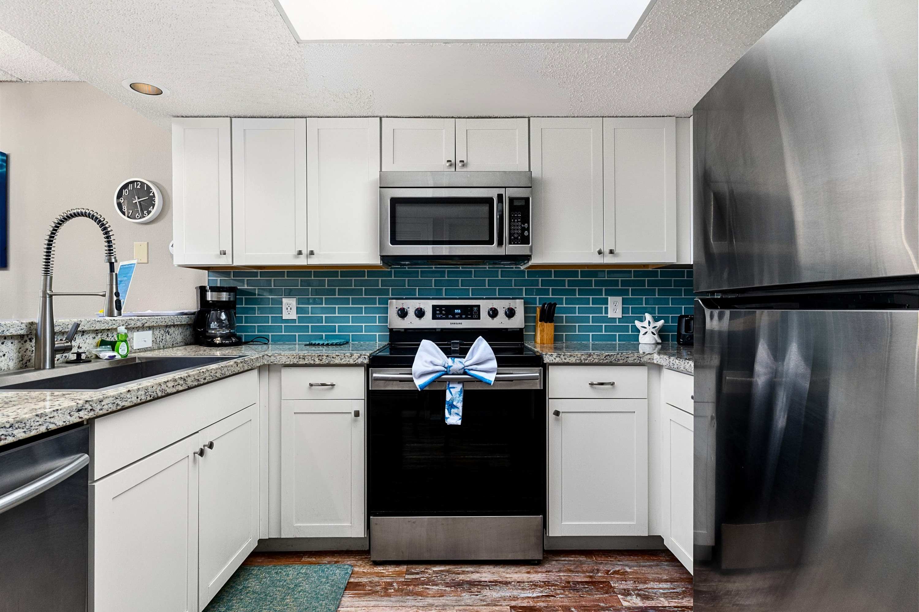 A close-up of the kitchen shows a stainless steel electric range with a black cooktop, a matching microwave above, and white shaker cabinets. A vibrant blue subway tile backsplash adds a pop of color, and a decorative bow is tied to the oven handle.