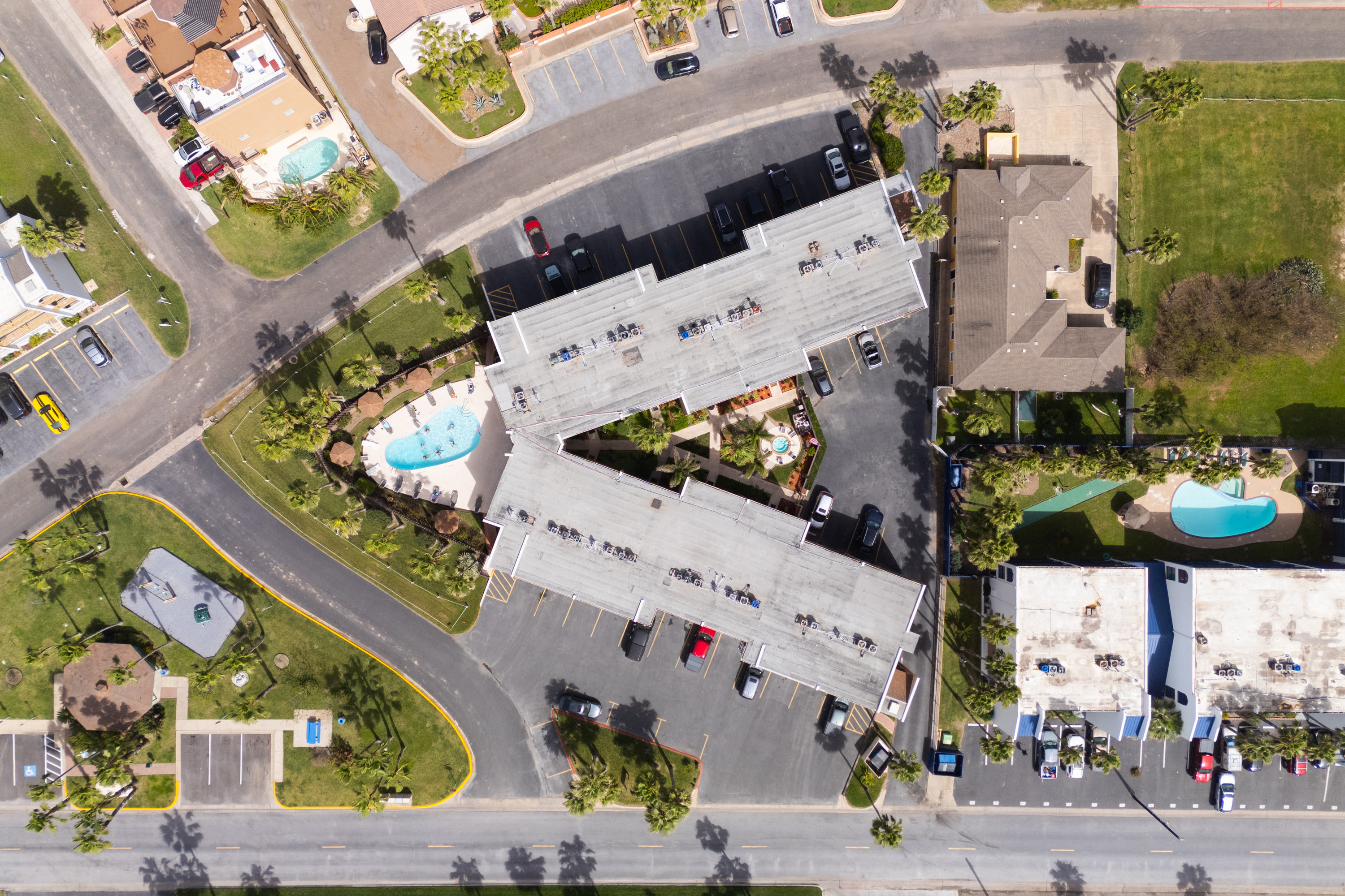 A closer aerial view of the condominium complex, highlighting the building layout, multiple swimming pools, and surrounding landscaping. Cars are visible in the parking areas.