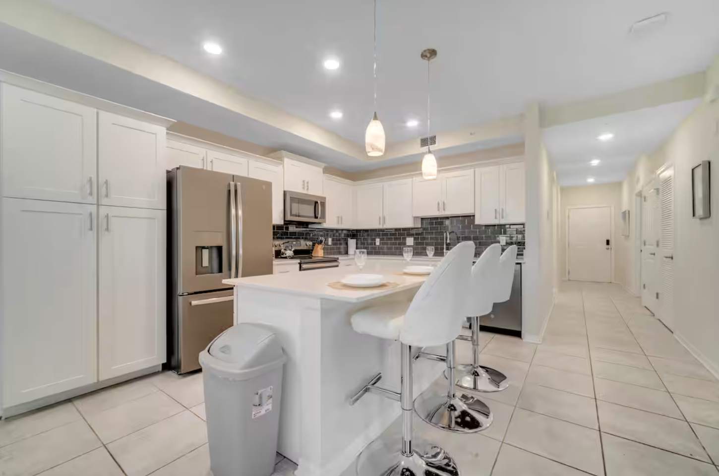 Wide shot of the kitchen with white shaker cabinets, stainless steel appliances, and a central island with two white bar stools. Recessed lighting and two pendant lights illuminate the space.