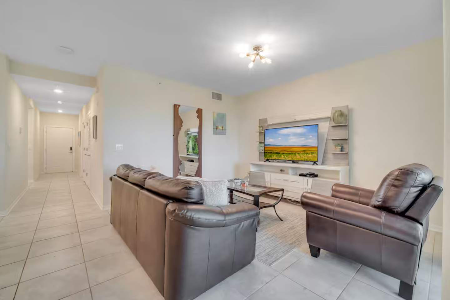 Inviting living room with a large flat-screen TV mounted on a white entertainment unit, a dark leather sofa, and light tile flooring, leading to a hallway and the balcony.