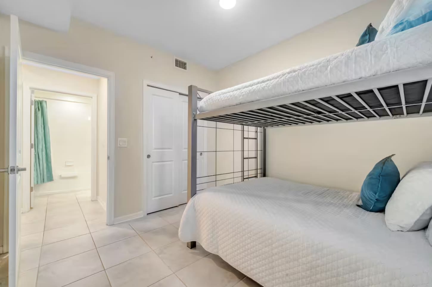 Another view of the bunk bed bedroom, showing a white sliding closet door and a doorway leading to an en-suite bathroom with a teal shower curtain visible. The room has light tiled floors.