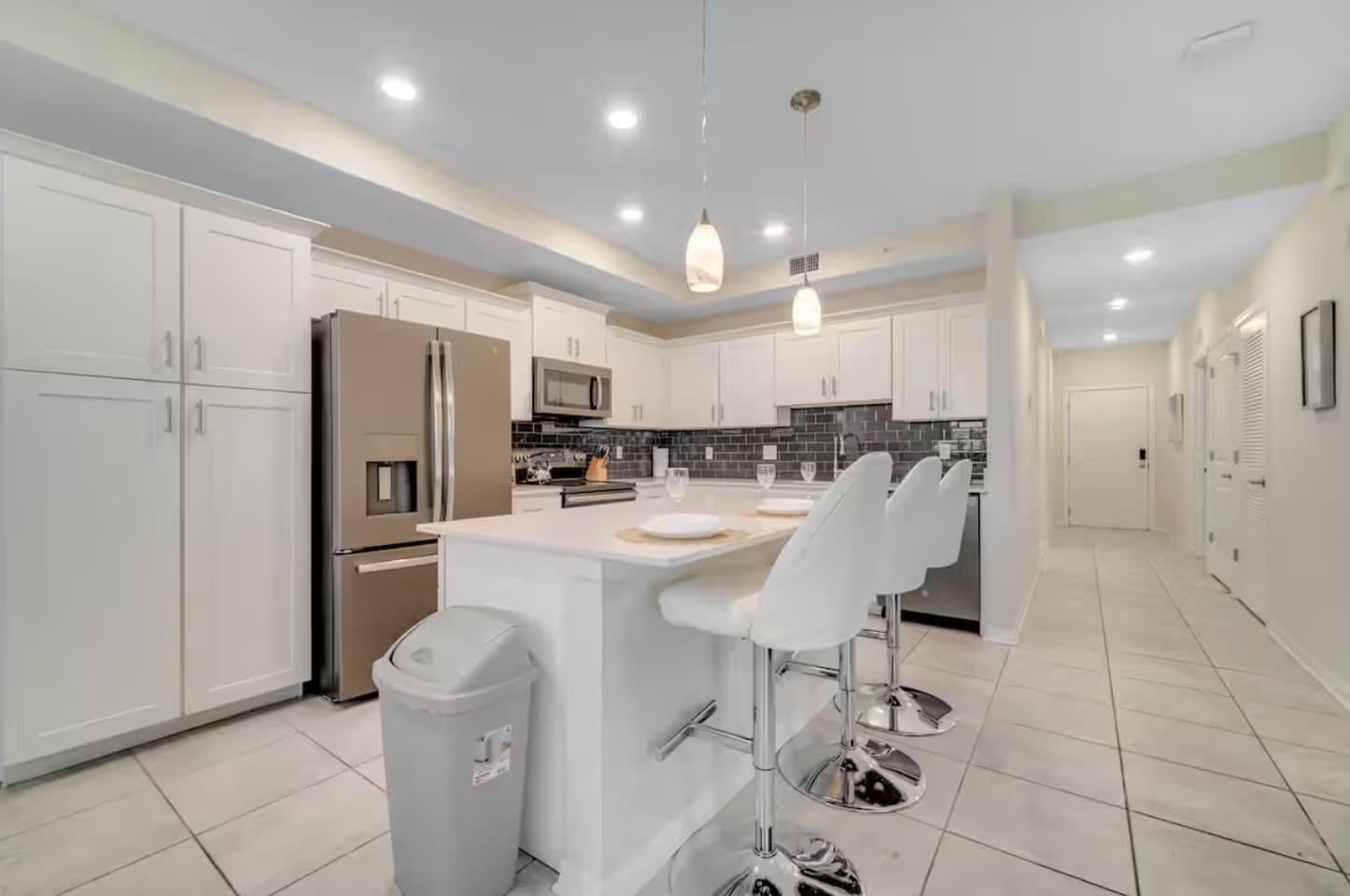 Wide shot of the kitchen with white shaker cabinets, stainless steel appliances, and a central island with two white bar stools. Recessed lighting and two pendant lights illuminate the space.