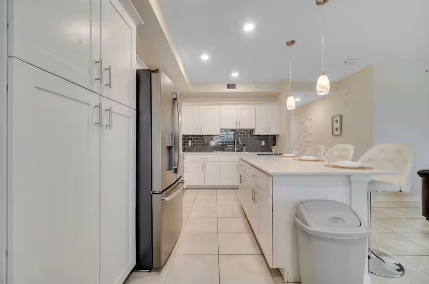 View of the kitchen from the hallway, featuring white shaker cabinets, stainless steel refrigerator, and a large island with pendant lighting. Light tile flooring extends throughout the space.