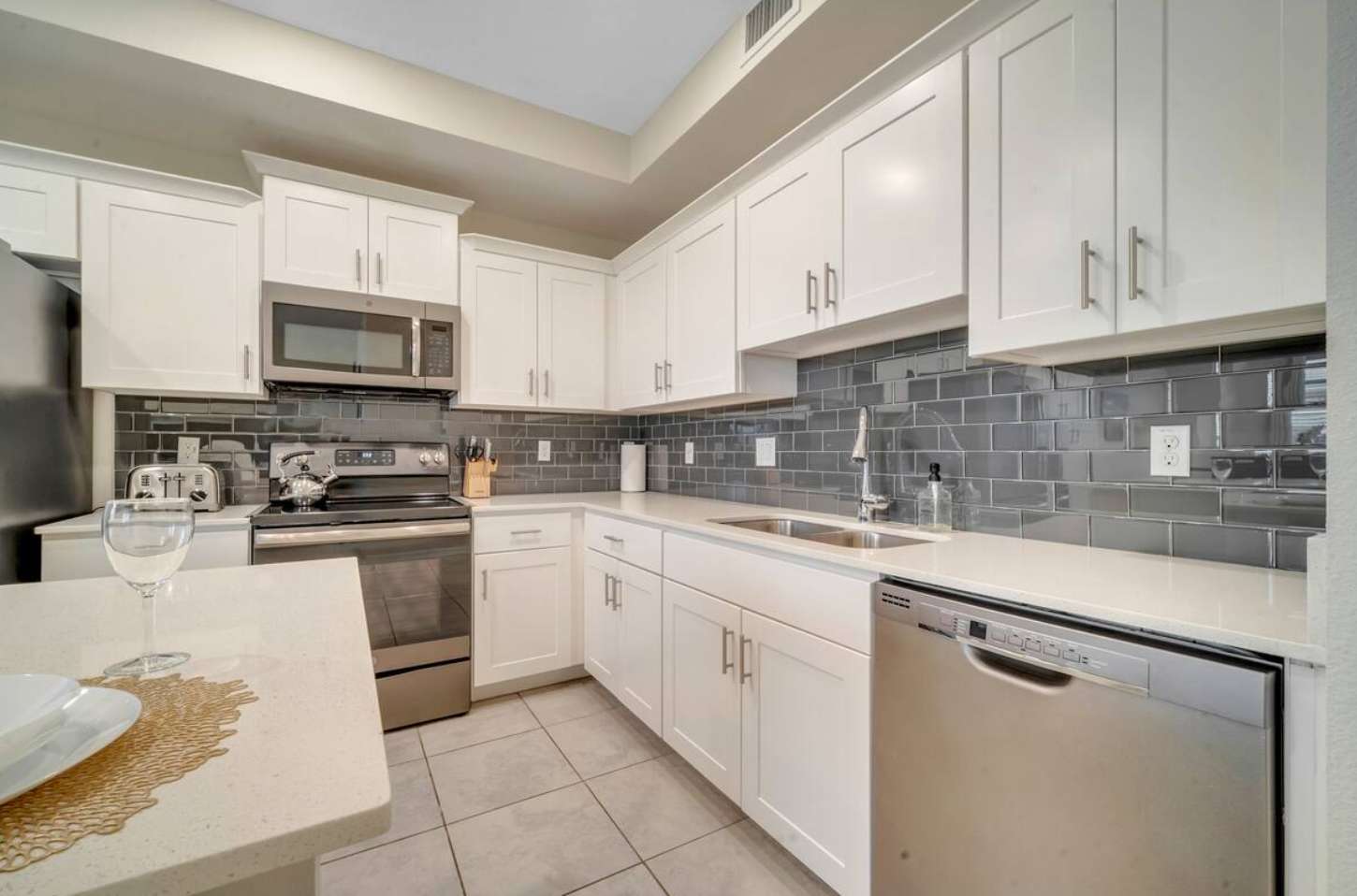 Modern kitchen with white shaker cabinets, light quartz countertops, and a dark gray subway tile backsplash. Stainless steel appliances include a microwave, range, and dishwasher.