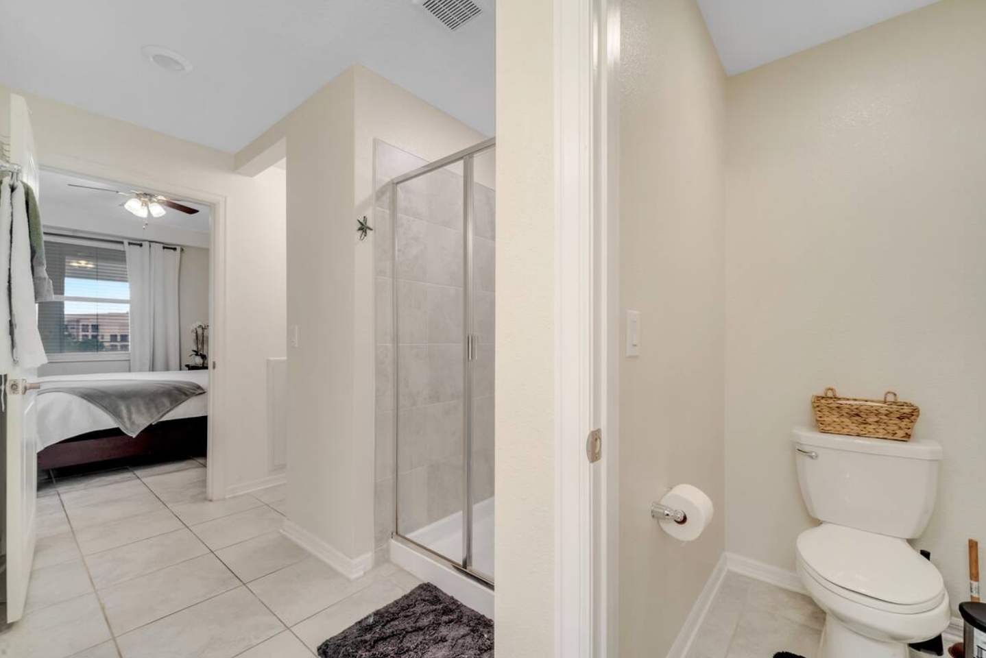 A light-colored bathroom featuring a glass-enclosed shower, a toilet, and a dark bath mat on light tiled floors. A doorway leads to a bedroom with a bed and ceiling fan visible in the background.