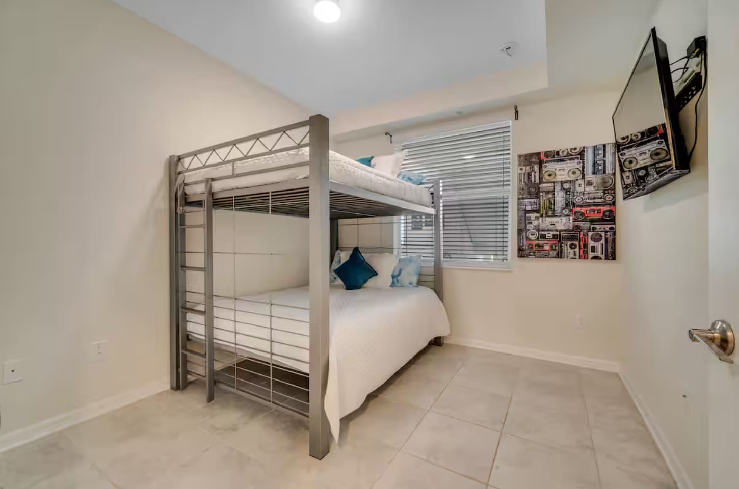 A cozy bedroom featuring a metal bunk bed with white bedding and blue accent pillows. A window with blinds and a colorful artwork depicting boomboxes adorn the wall, with a wall-mounted TV nearby.