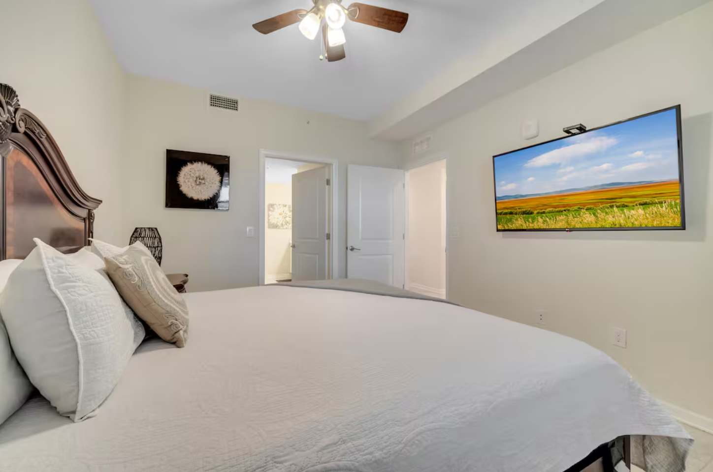 Master bedroom featuring a large dark wood headboard, white bedding, and a wall-mounted flat-screen TV. A ceiling fan with light fixture is centered above the bed.