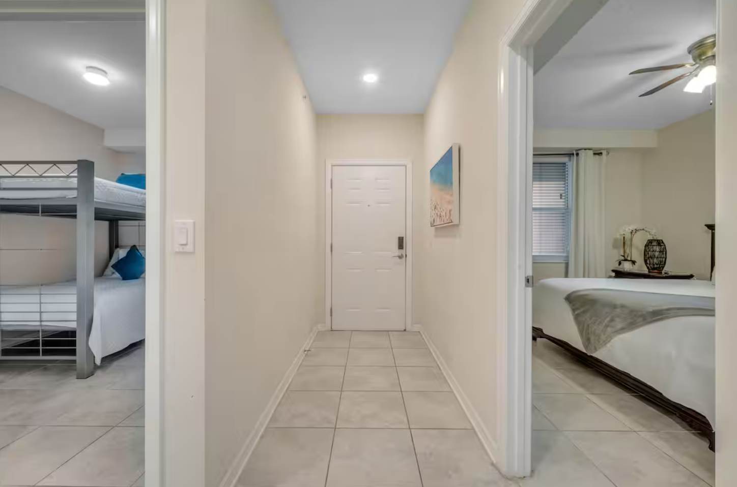 A bright hallway with light tiled floors and cream-colored walls, leading to a white front door. Doorways on either side open into two separate bedrooms, one with bunk beds and the other with a standard bed.
