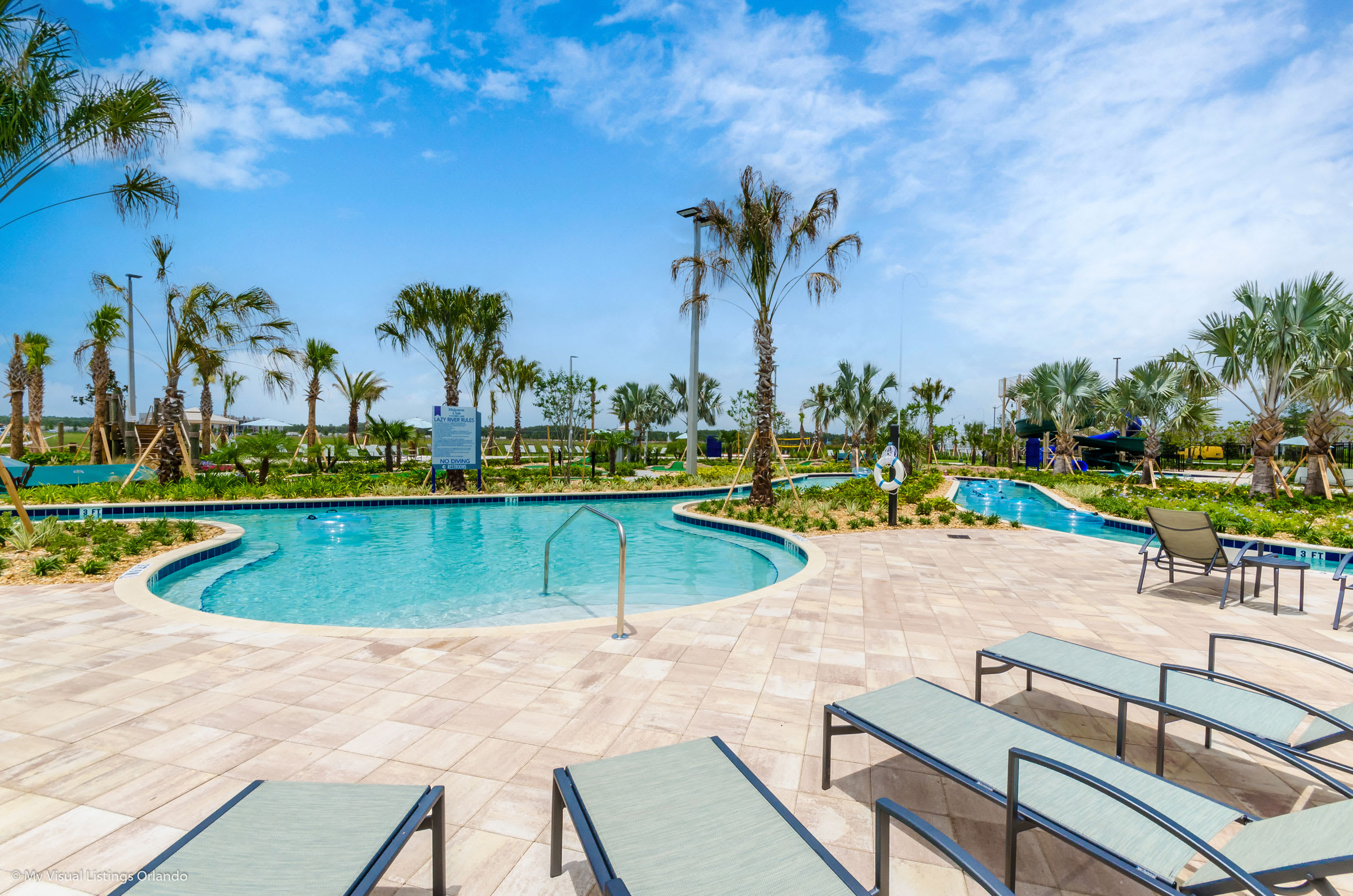 Resort-style swimming pool with a shallow entry area and surrounding lounge chairs on a light-colored paved deck. Palm trees and a multi-story water slide structure are visible in the background under a bright blue sky.