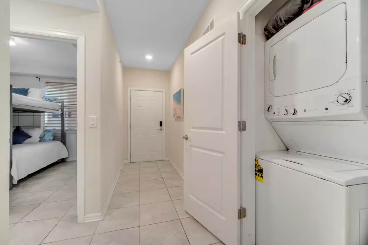 A hallway view showing a stacked white washer and dryer unit neatly tucked into a closet with a white door. The hallway also provides a glimpse into a bedroom with bunk beds.