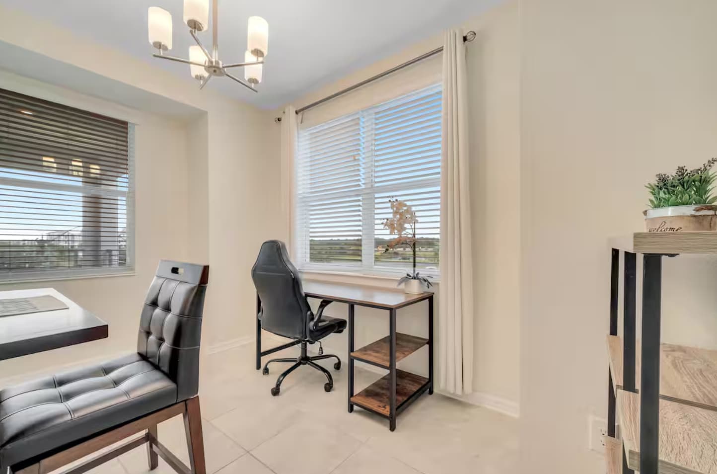 Bright home office space featuring a black ergonomic office chair, a modern wooden desk with metal shelving, and a large window with white blinds overlooking a green landscape. A contemporary chandelier hangs from the ceiling.