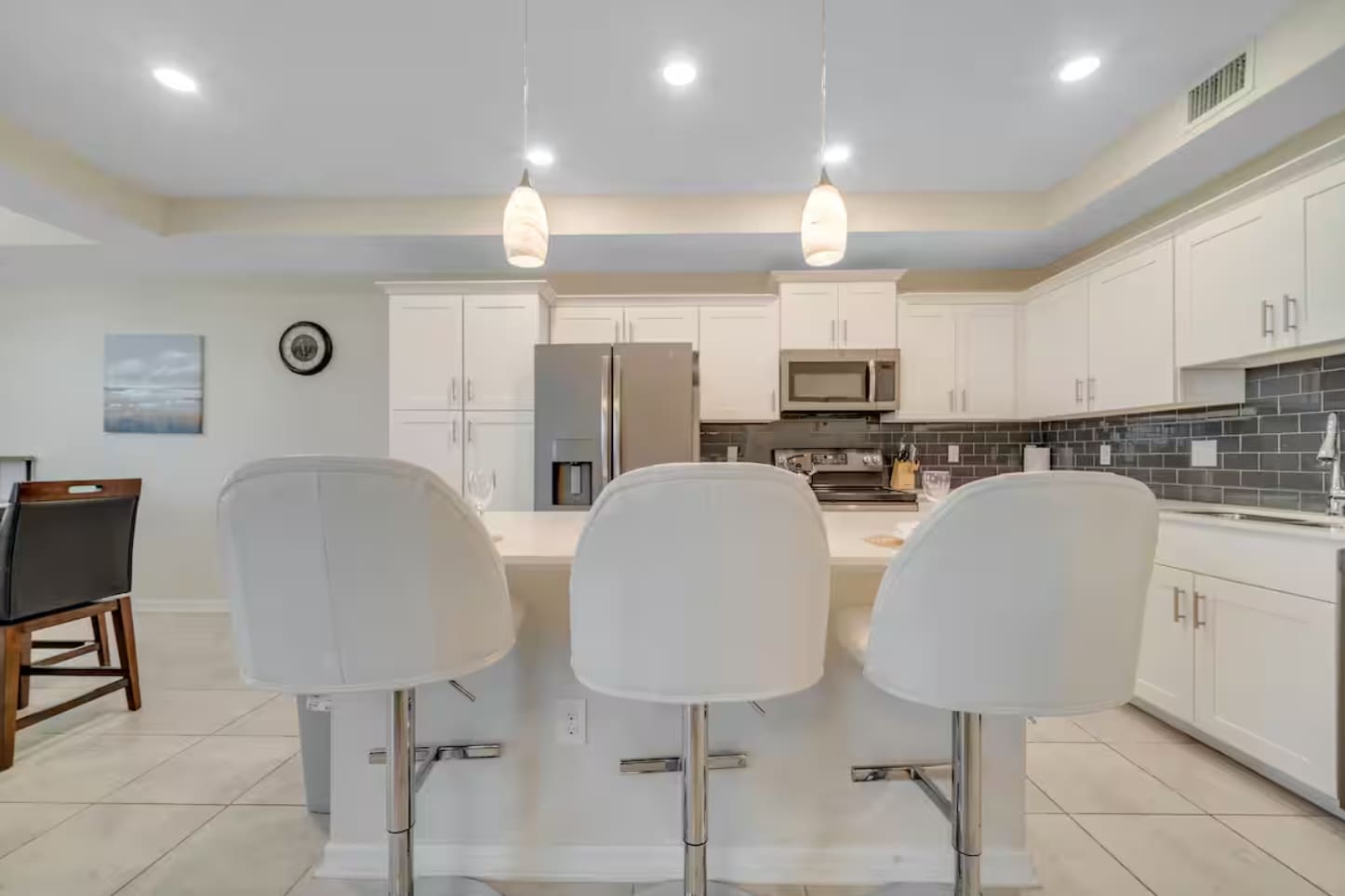 View of the kitchen island with three white bar stools, looking towards the main kitchen area with white cabinets and stainless steel appliances. Two pendant lights hang above the island.